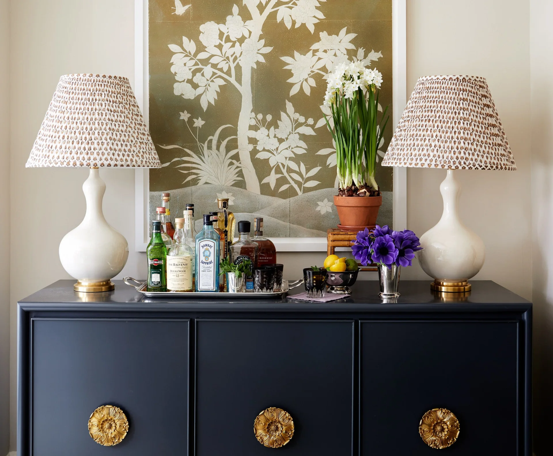 A navy blue console table with floral brass hardware is styled with bottles, two white table lamps with patterned shades, and botanical art.