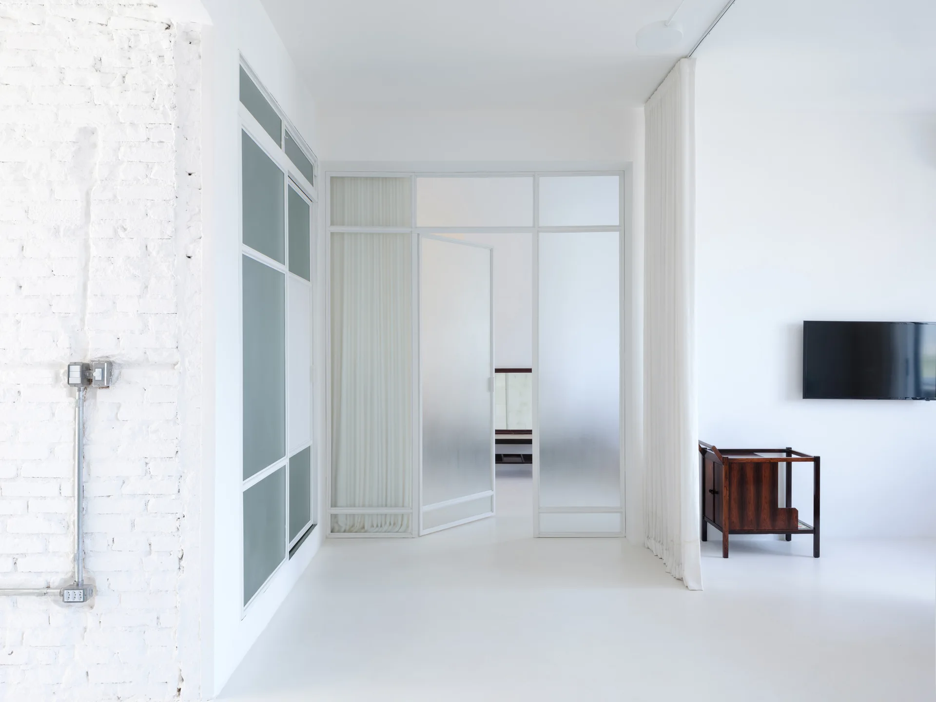 A minimalist white room features a painted brick wall, a white-framed frosted glass partition door, a small dark wood cabinet, and a wall-mounted TV.