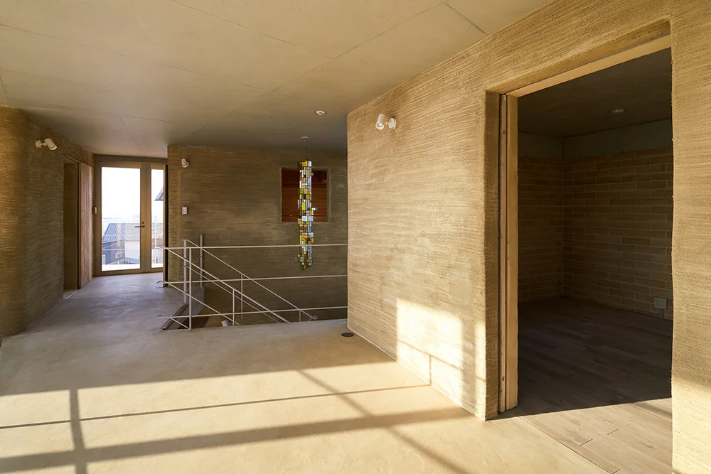 An interior hallway with rough tan plaster walls, a smooth light grey ceiling, and a thin metal staircase railing.