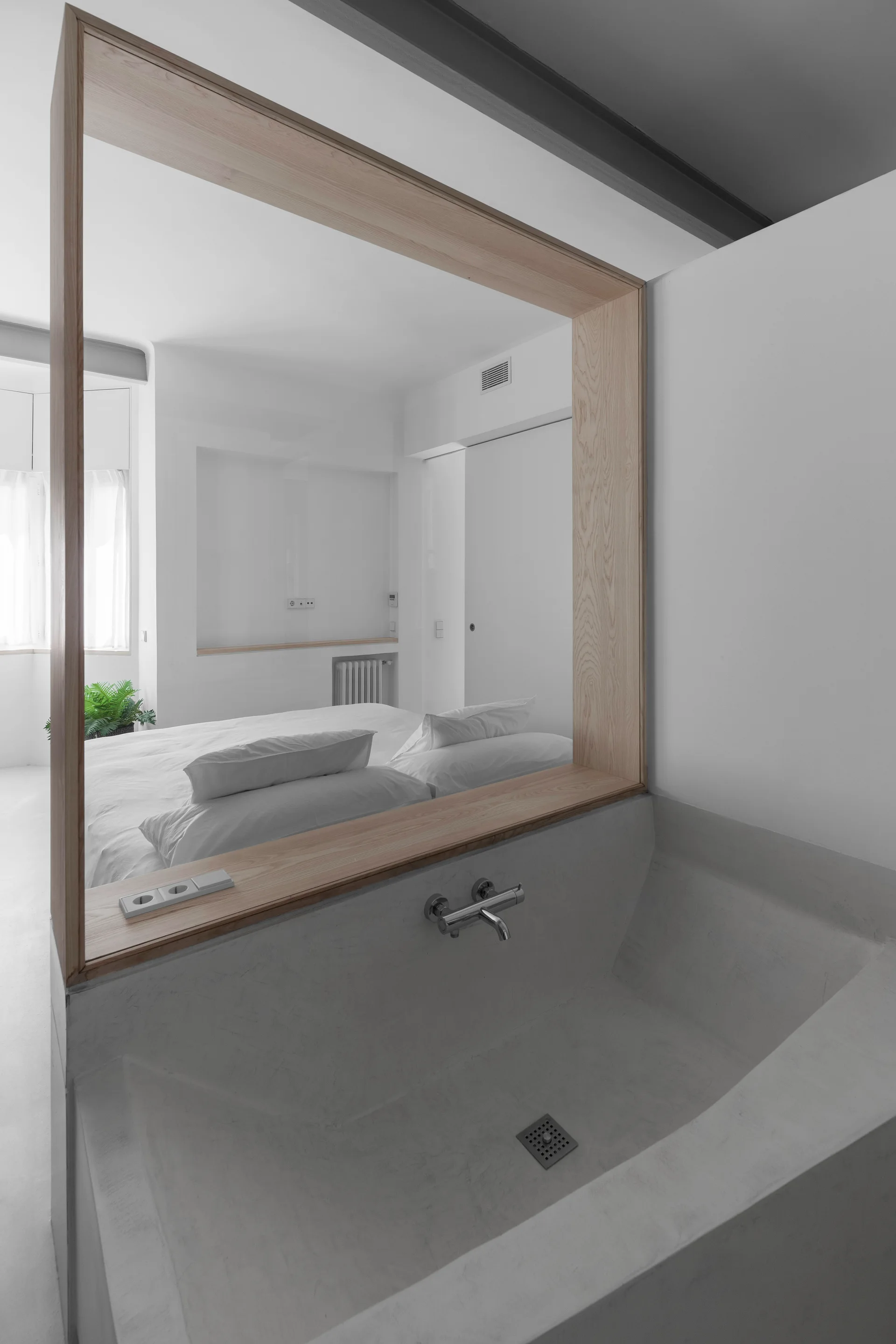 A minimalist bathroom featuring a grey concrete basin and a wall-mounted faucet below a large wood-framed opening looking into a white bedroom.