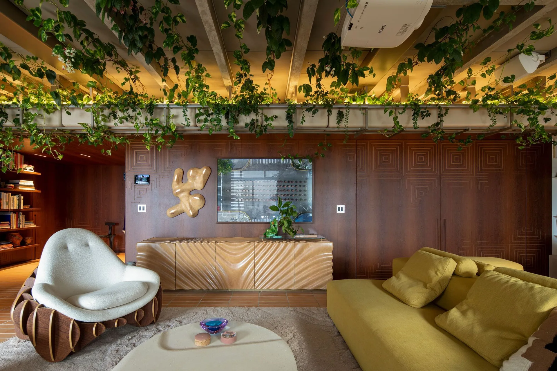 A living room with a yellow sofa, sculptural armchair, wavy wood sideboard, and hanging plants draping from the exposed concrete ceiling beams.