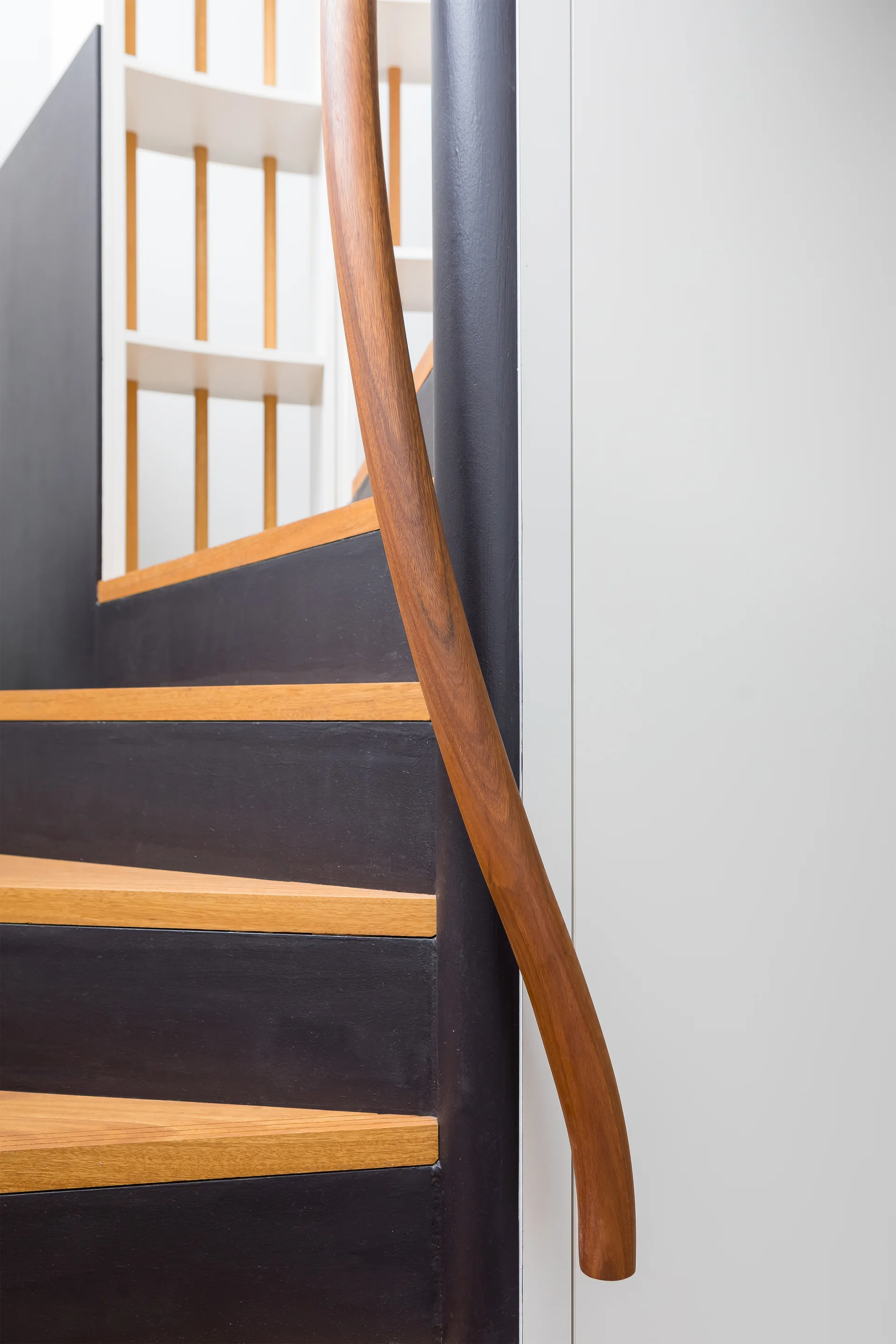 A close-up of a modern staircase featuring light wood treads, matte black risers, and a curved wooden handrail against a white wall.