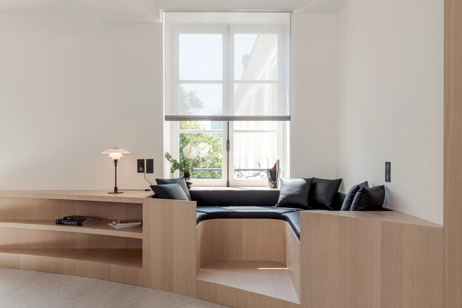 A curved light wood built-in bench with black cushions sits below a window, flanked by integrated wooden shelving and a mushroom-shaped table lamp.