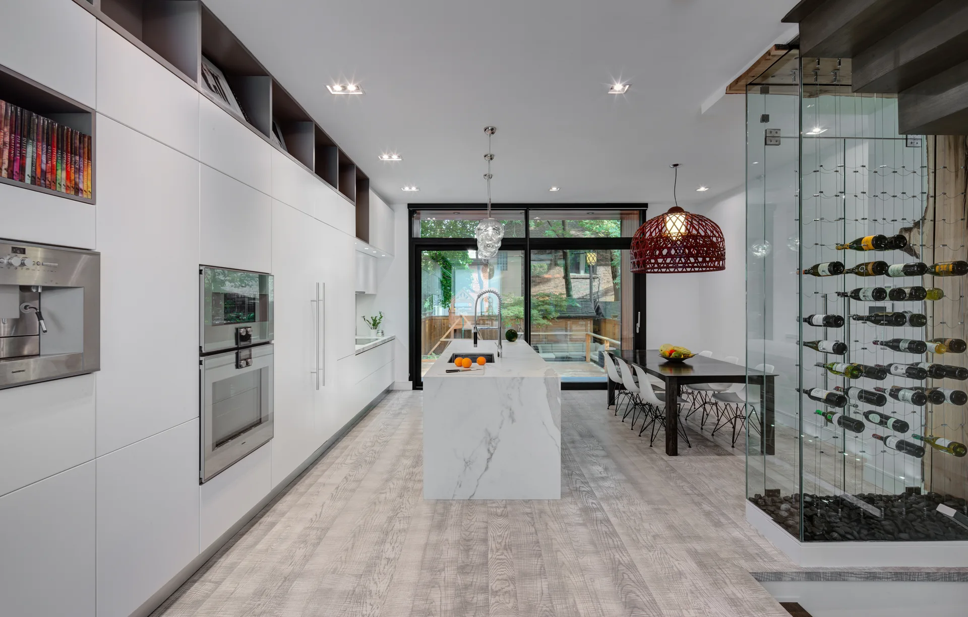 A modern kitchen with white seamless cabinets, a central marble island, and a glass-enclosed wine storage unit beside a dining table.