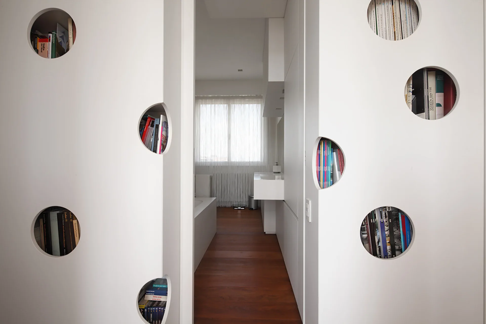 A view through a white partition wall with circular cutout bookshelves looking into a minimalist bathroom with dark wood flooring.