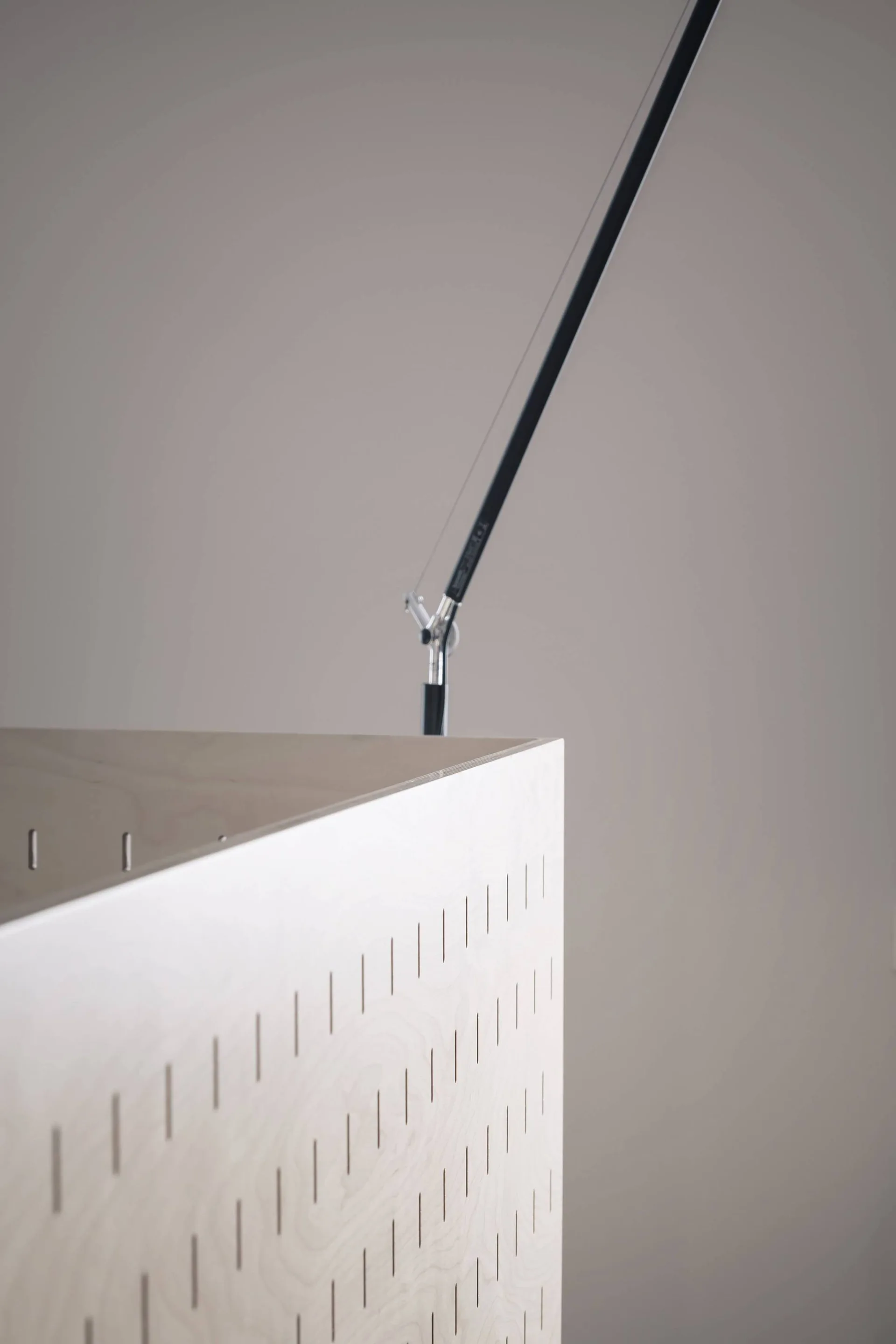 A close-up of a light wood plywood cabinet featuring diagonal slotted cutouts next to a slim black metal lamp against a neutral wall.