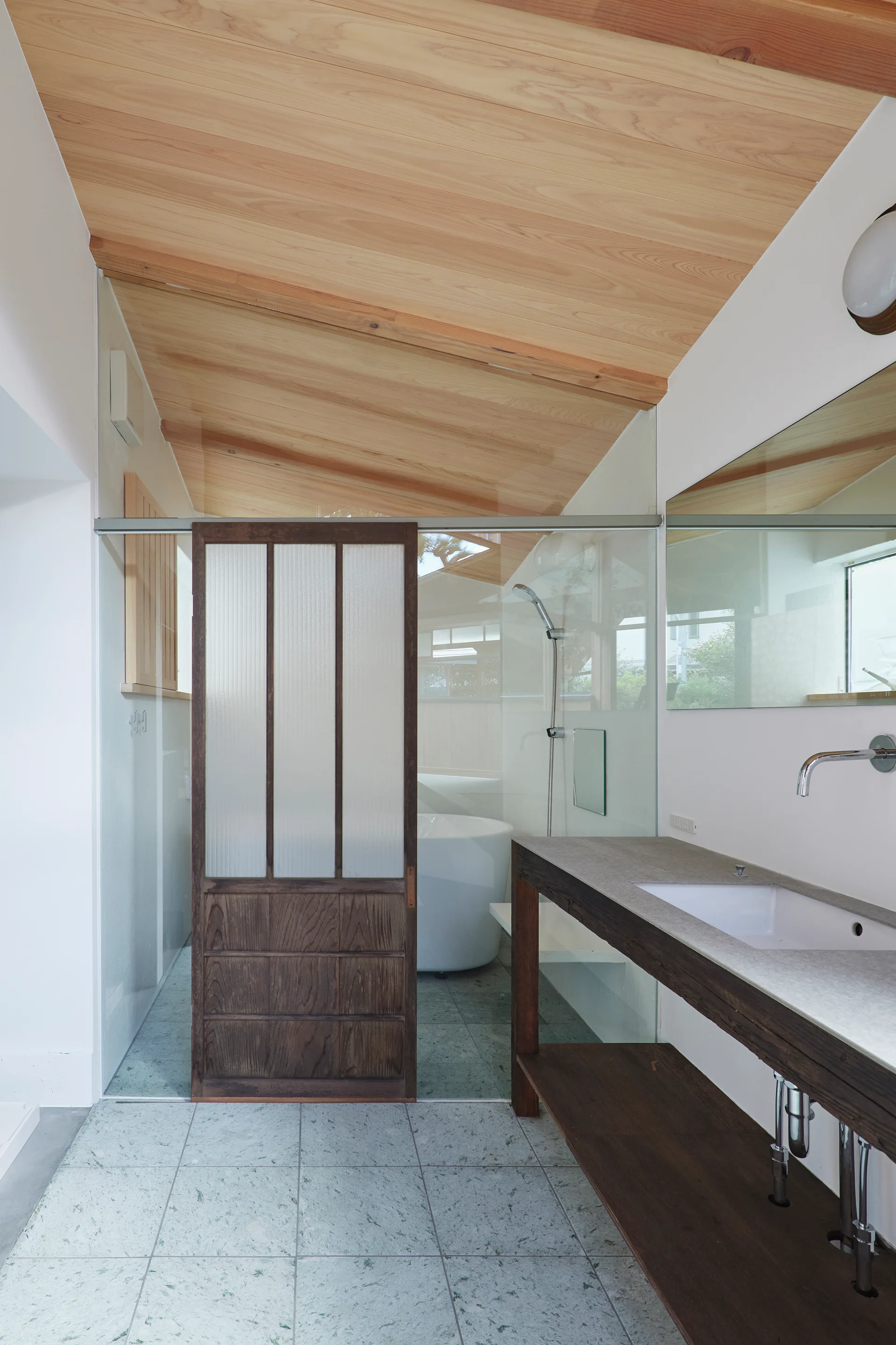A minimalist bathroom featuring a sloped light wood ceiling, a dark wood vanity, speckled grey floor tiles, and a glass-enclosed wet room.
