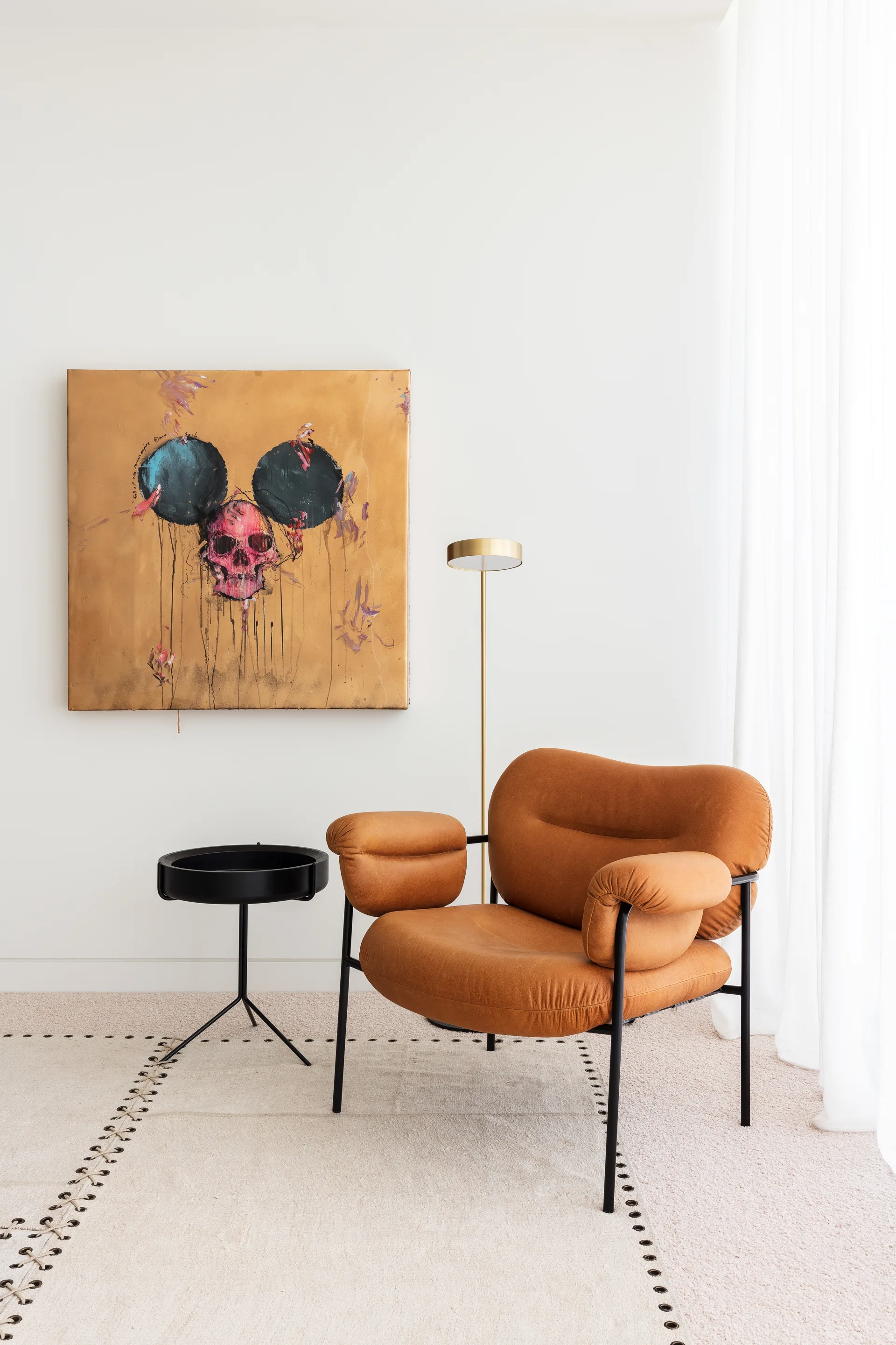 A tan leather armchair sits next to a black side table and slim gold floor lamp below a large gold painting on a white wall.