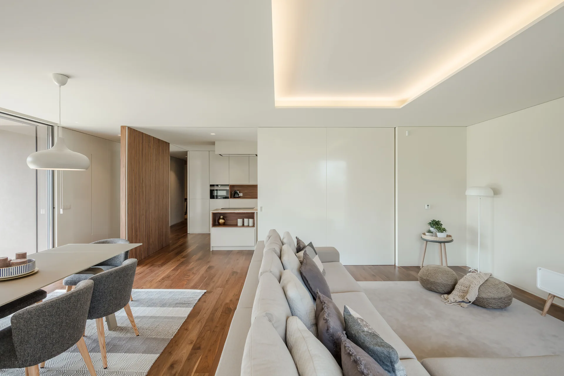 A minimalist living room features a cream modular sofa, a vertical wood slat partition, and a dining table under a modern pendant light.