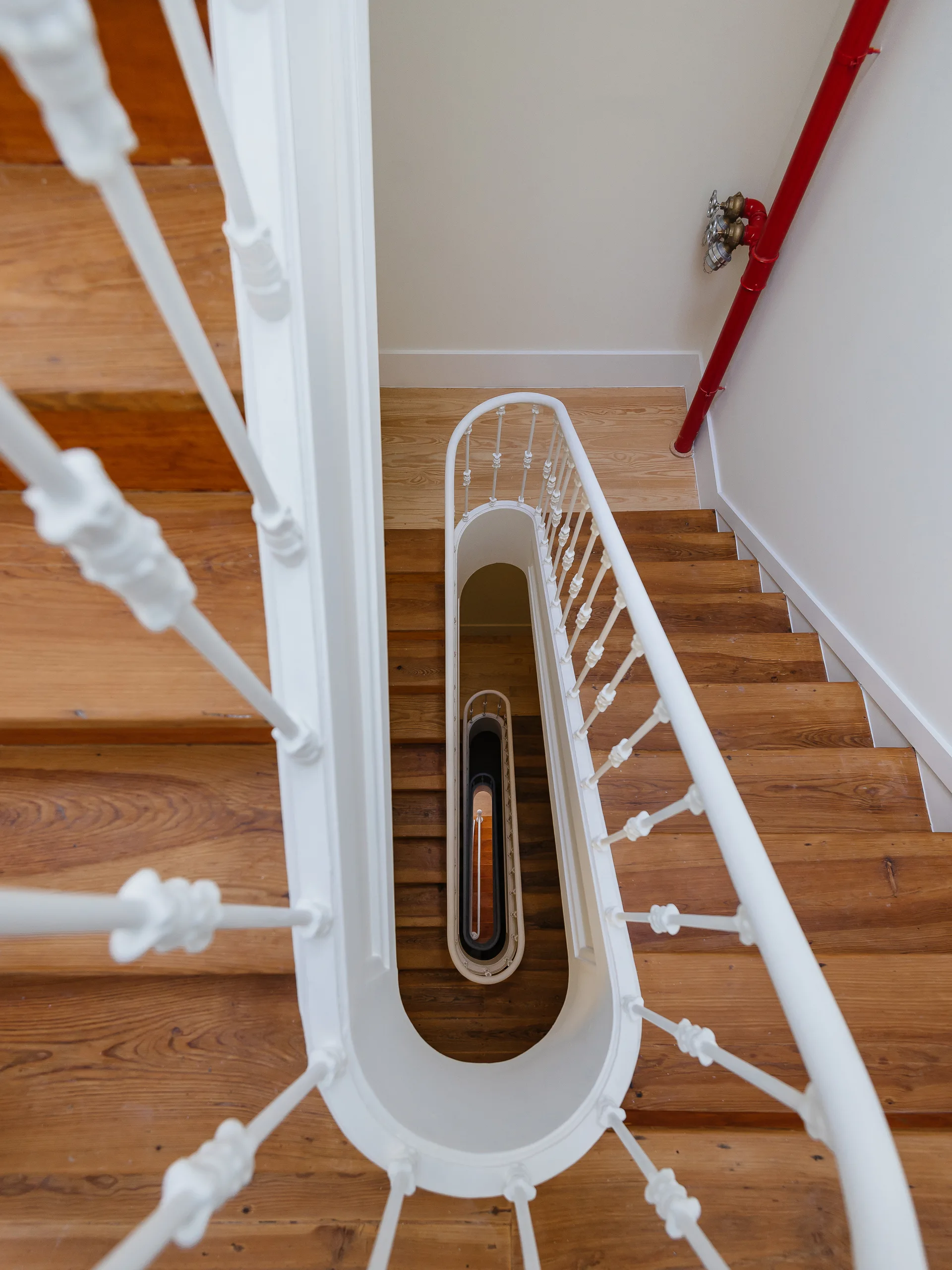 Looking down a spiral wooden staircase with a white decorative balustrade and a bright red utility pipe on the adjacent white wall.