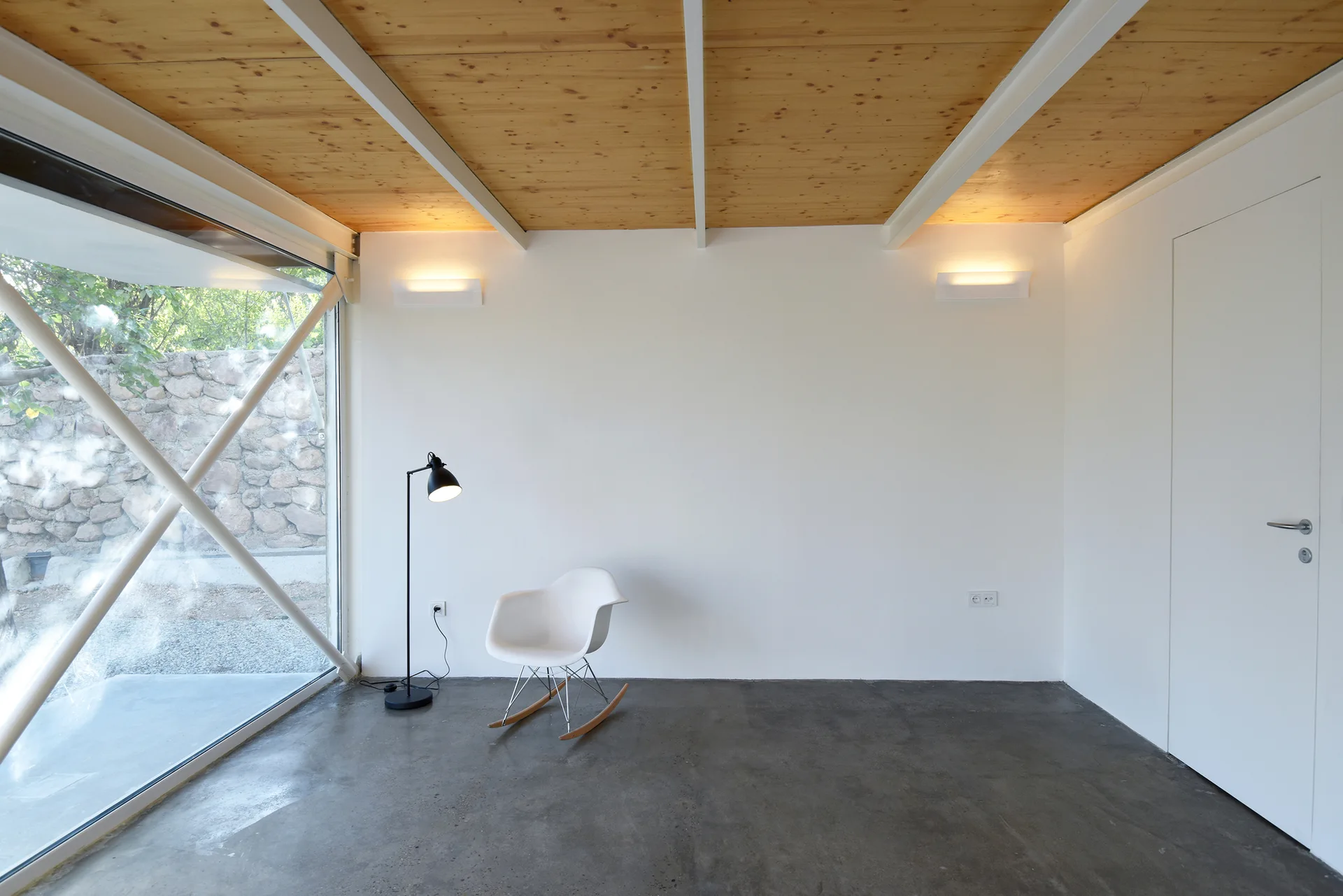A minimalist room with a white shell rocking chair, a black floor lamp, smooth concrete flooring, and a natural wood plank ceiling.