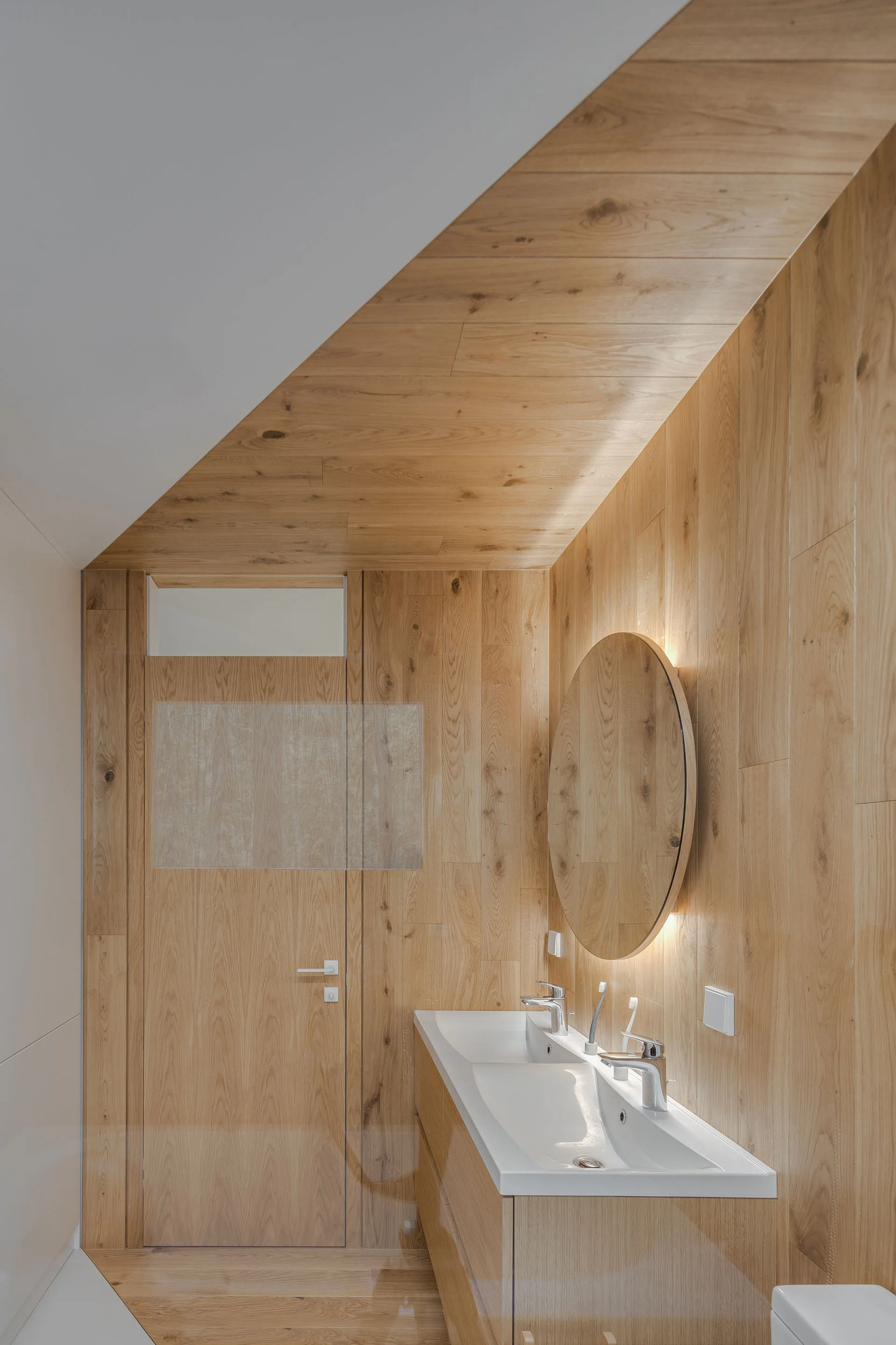 A modern bathroom features warm wood paneling across the walls and ceiling, a round backlit mirror, and a sleek white double sink vanity.