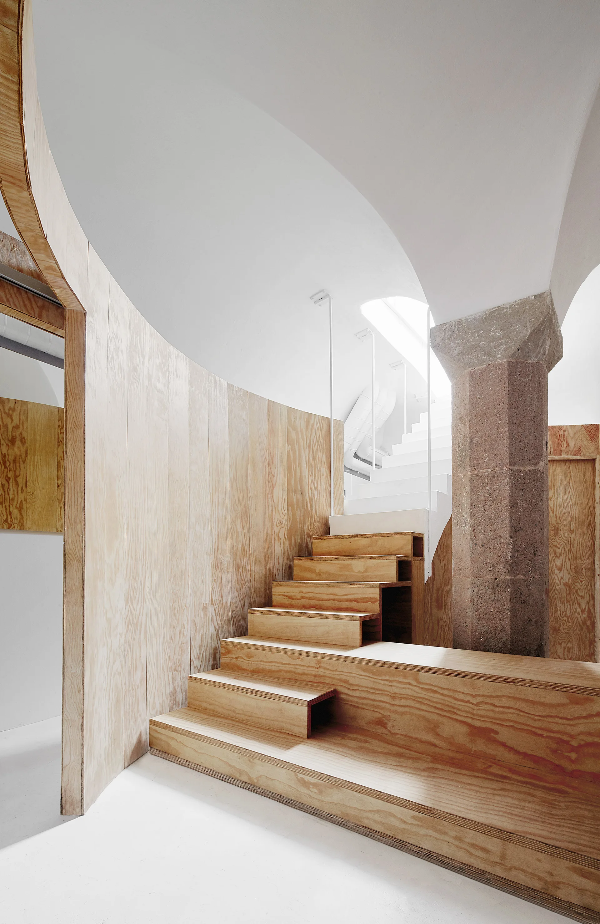 A modern plywood staircase winds upward next to a historic octagonal stone column beneath a white vaulted ceiling.