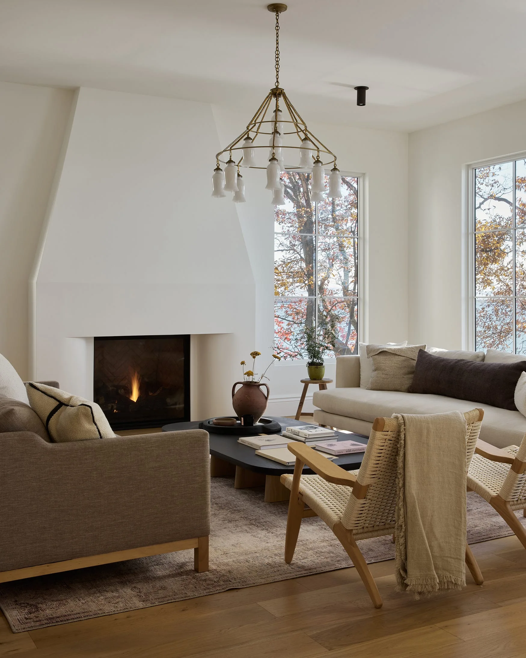 A living room features a white plaster fireplace, a black coffee table, two woven armchairs, and large grid-frame windows with a view of autumn trees.