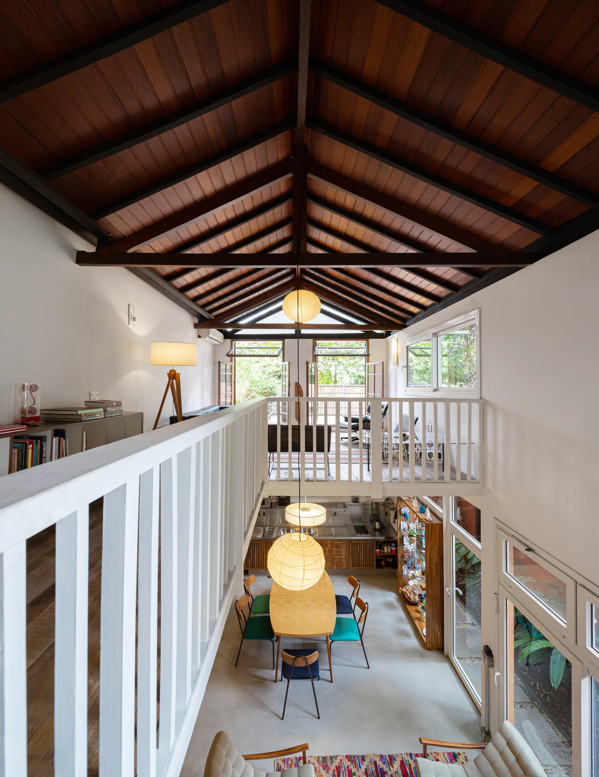 A high-angle view of a loft dining area with a light wood table, paper pendant lights, and a white mezzanine railing under a vaulted wood ceiling.