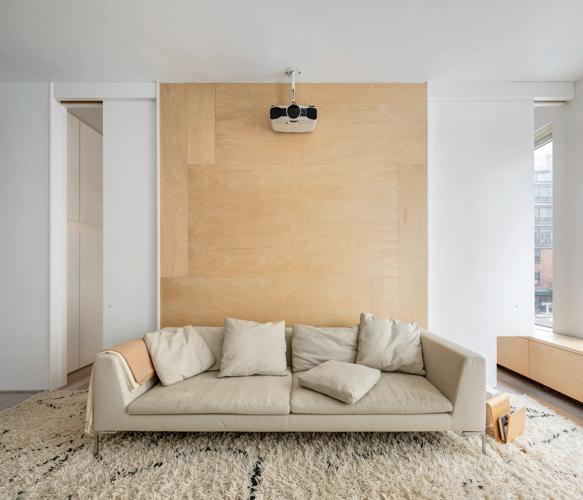 A modern beige fabric sofa on a thick white shag rug in front of a light plywood feature wall with a mounted projector.
