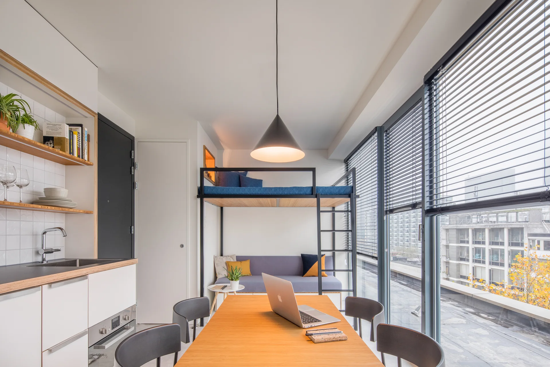 A modern studio apartment featuring a black metal loft bed over a grey sofa, adjacent to a wooden dining table and a white kitchenette.