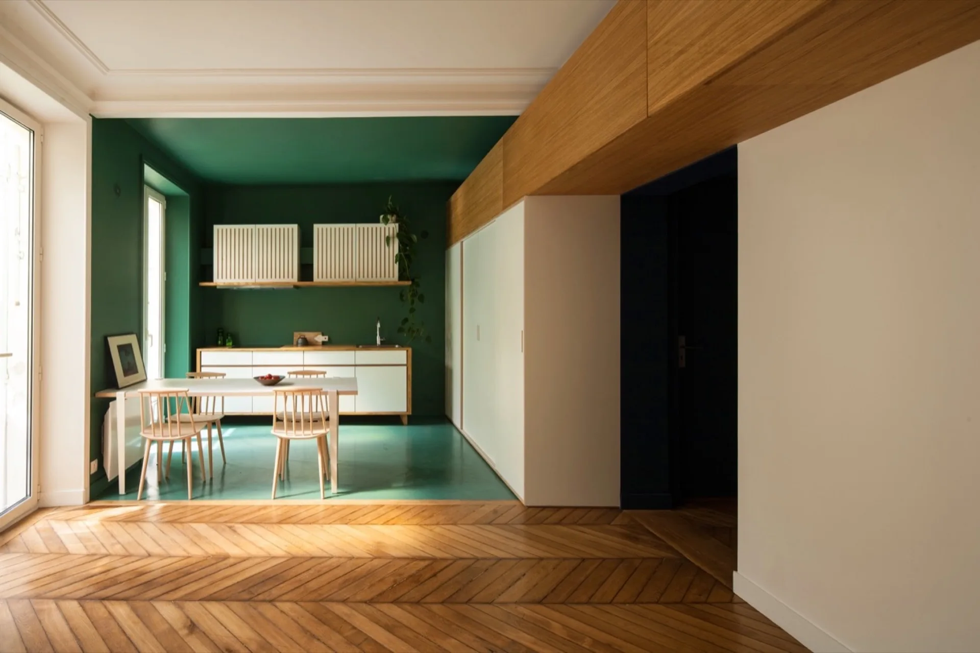 A modern kitchen with deep green walls and ceiling features white cabinets, a light wood dining table with spindle chairs, and parquet flooring.