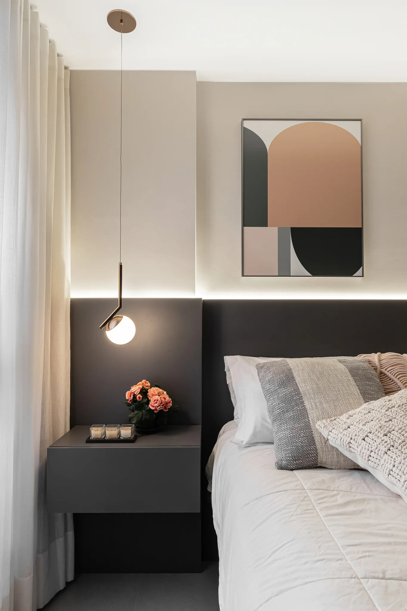 A modern bedroom features a floating gray nightstand, a backlit dark headboard, a sphere pendant light, and geometric wall art.