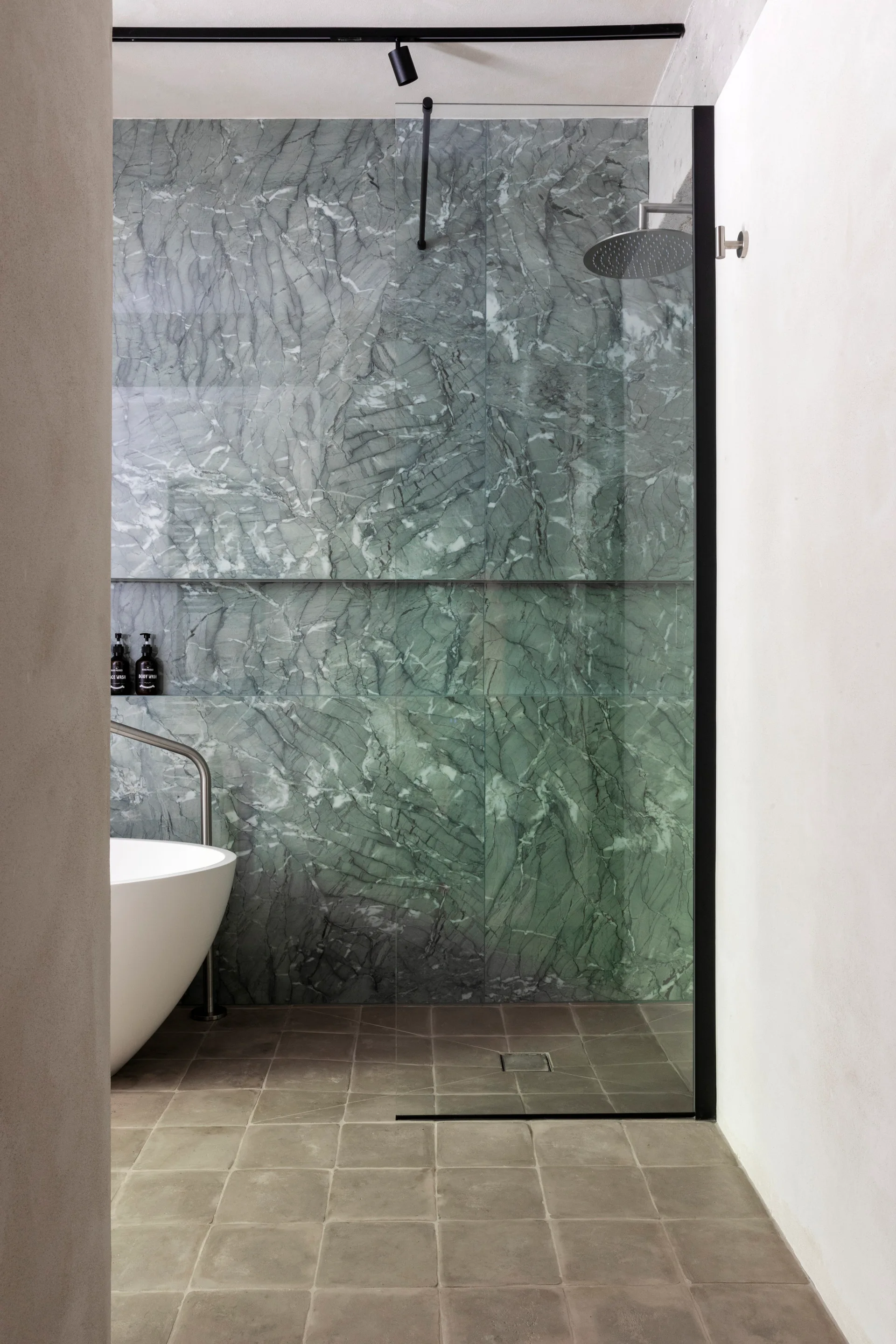 A modern bathroom features a green veined marble tiled shower wall with a recessed shelf, next to a white freestanding tub and grey floor tiles.