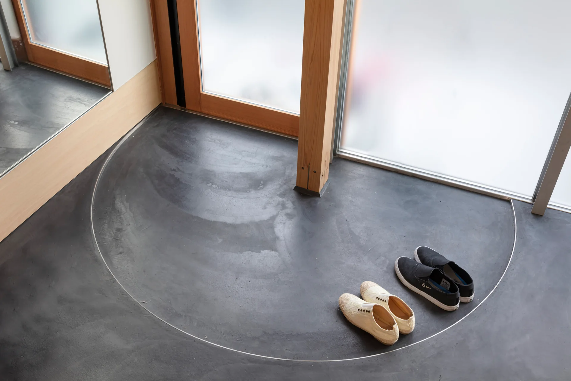 A close-up view of a modern entry floor with a dark concrete finish, a curved metal inlay, wood door frames, and two pairs of shoes.