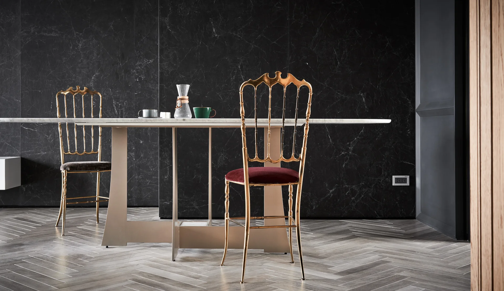 Ornate gold metal dining chairs are arranged around a white marble table against a dark marble wall with grey herringbone wood flooring.