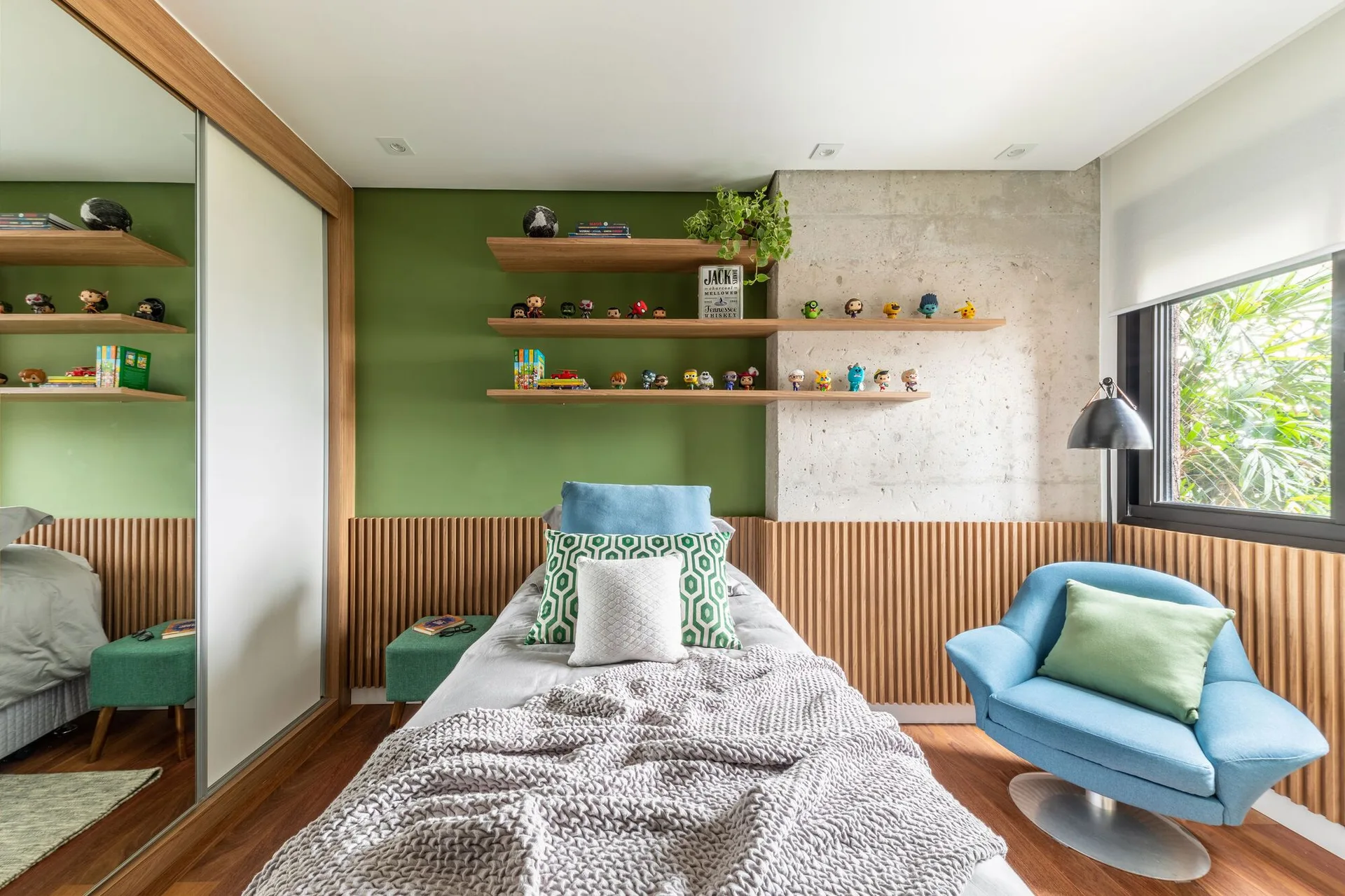 A modern bedroom with a bed set against a green and concrete wall featuring lower wood slat paneling, floating shelves, and a blue armchair.