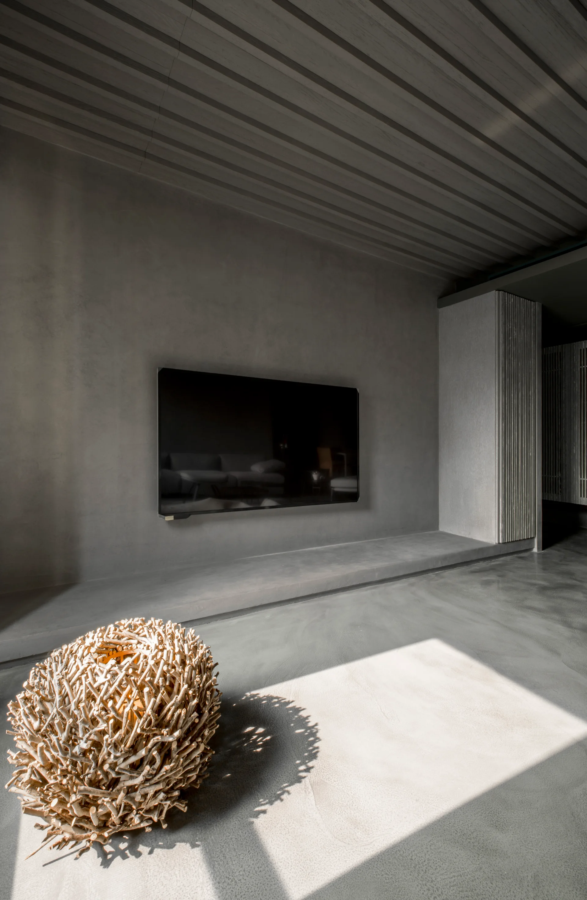 A wall-mounted television hangs on a grey plaster wall above a polished concrete floor, with a decorative twig sphere resting in a patch of sunlight.