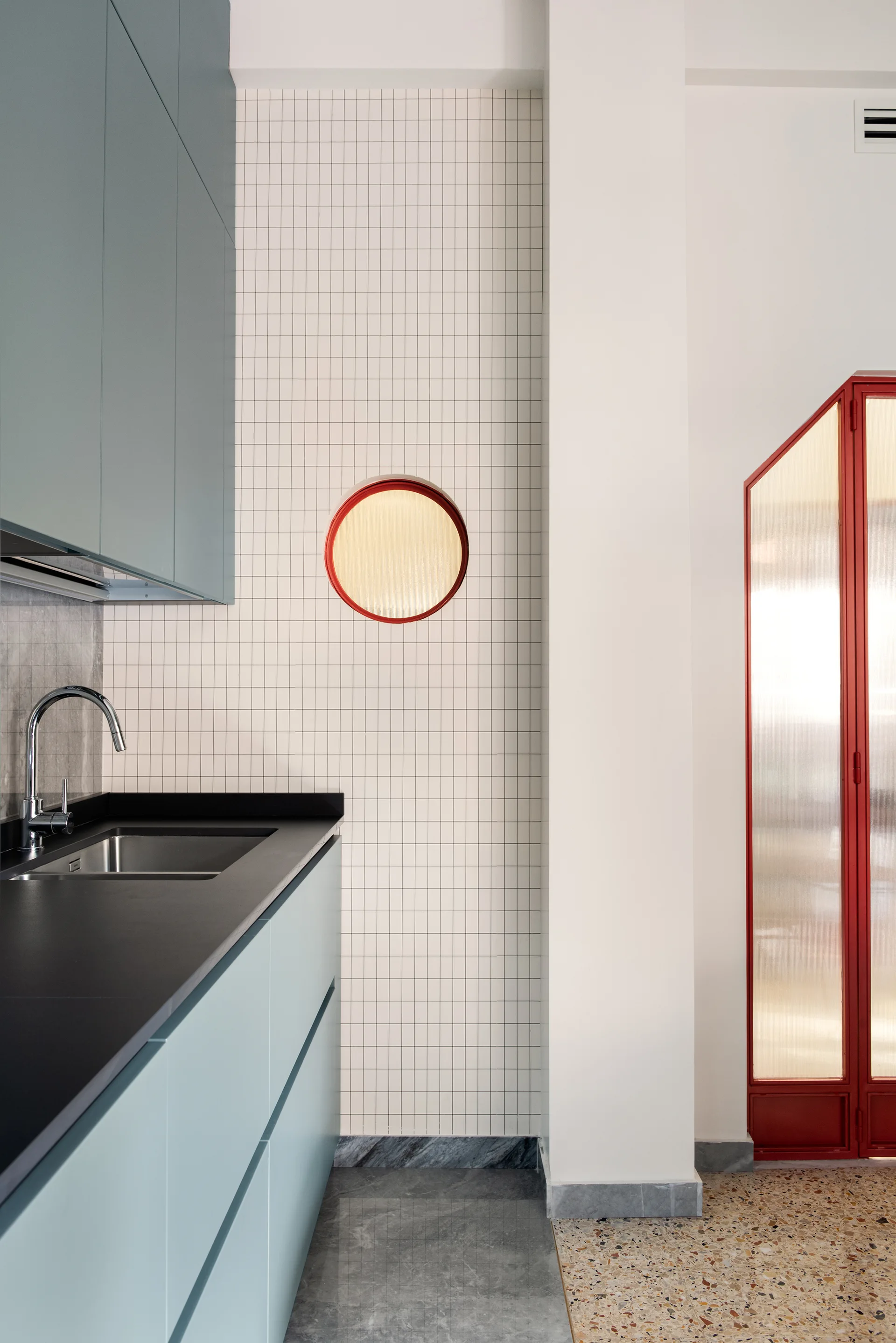 A modern kitchen features matte blue-green cabinetry, a white grid tile backsplash with a circular red-framed window, and polished terrazzo flooring.