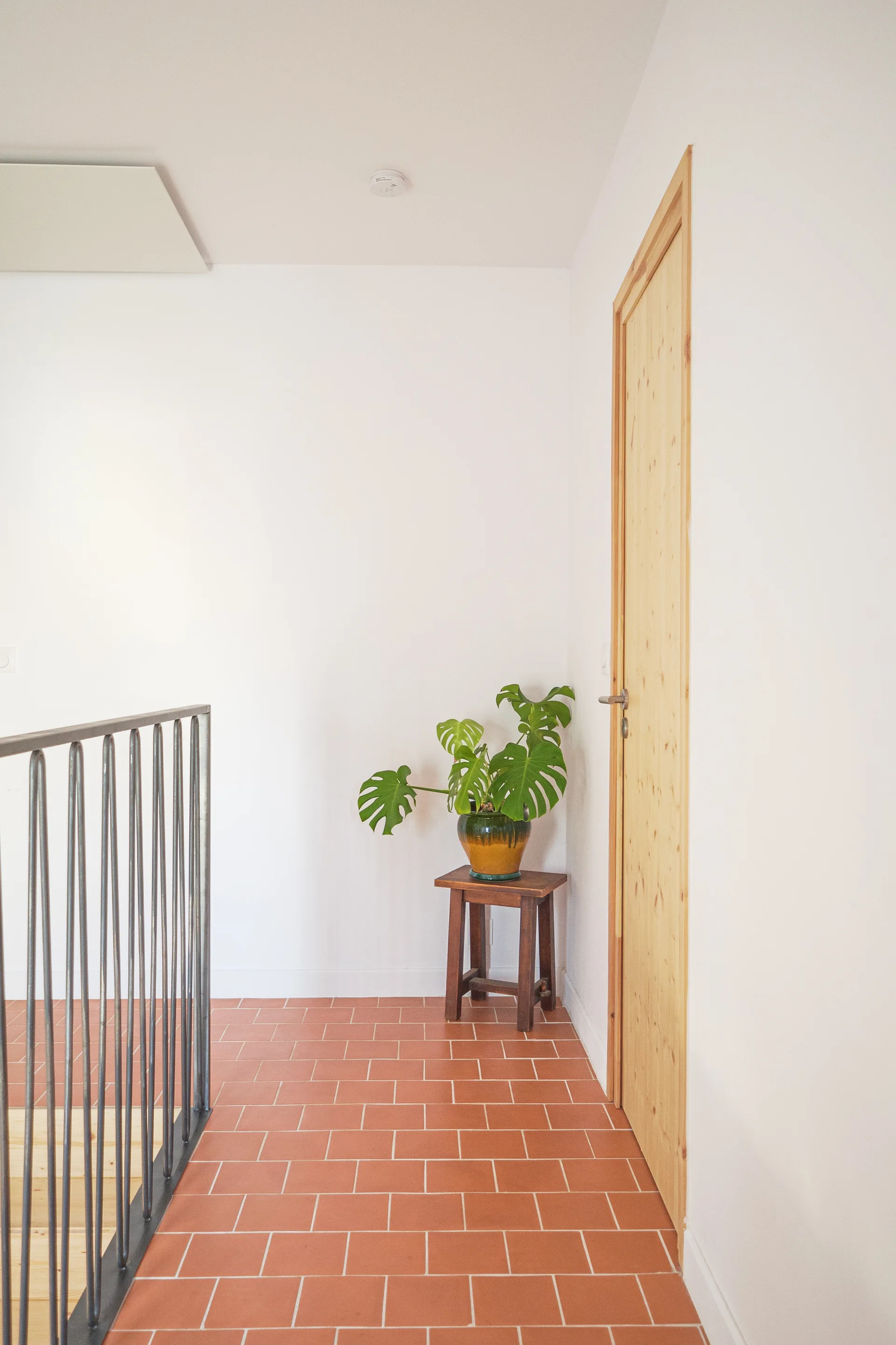 A bright staircase landing features terracotta tile flooring, a natural wood door, a black metal railing, and a potted monstera plant on a stool.