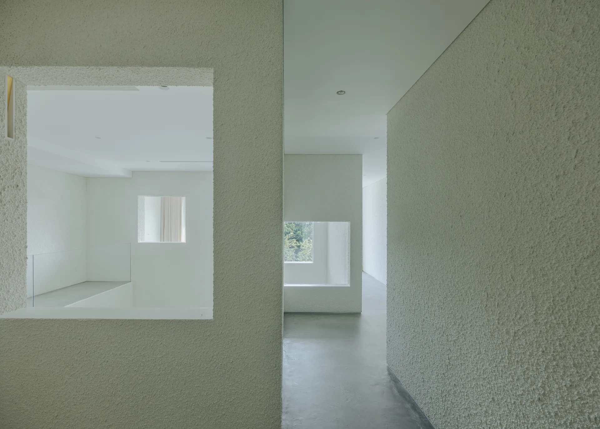 A minimalist corridor with highly textured plaster walls features rectangular cutouts looking into adjacent spaces over a smooth concrete floor.