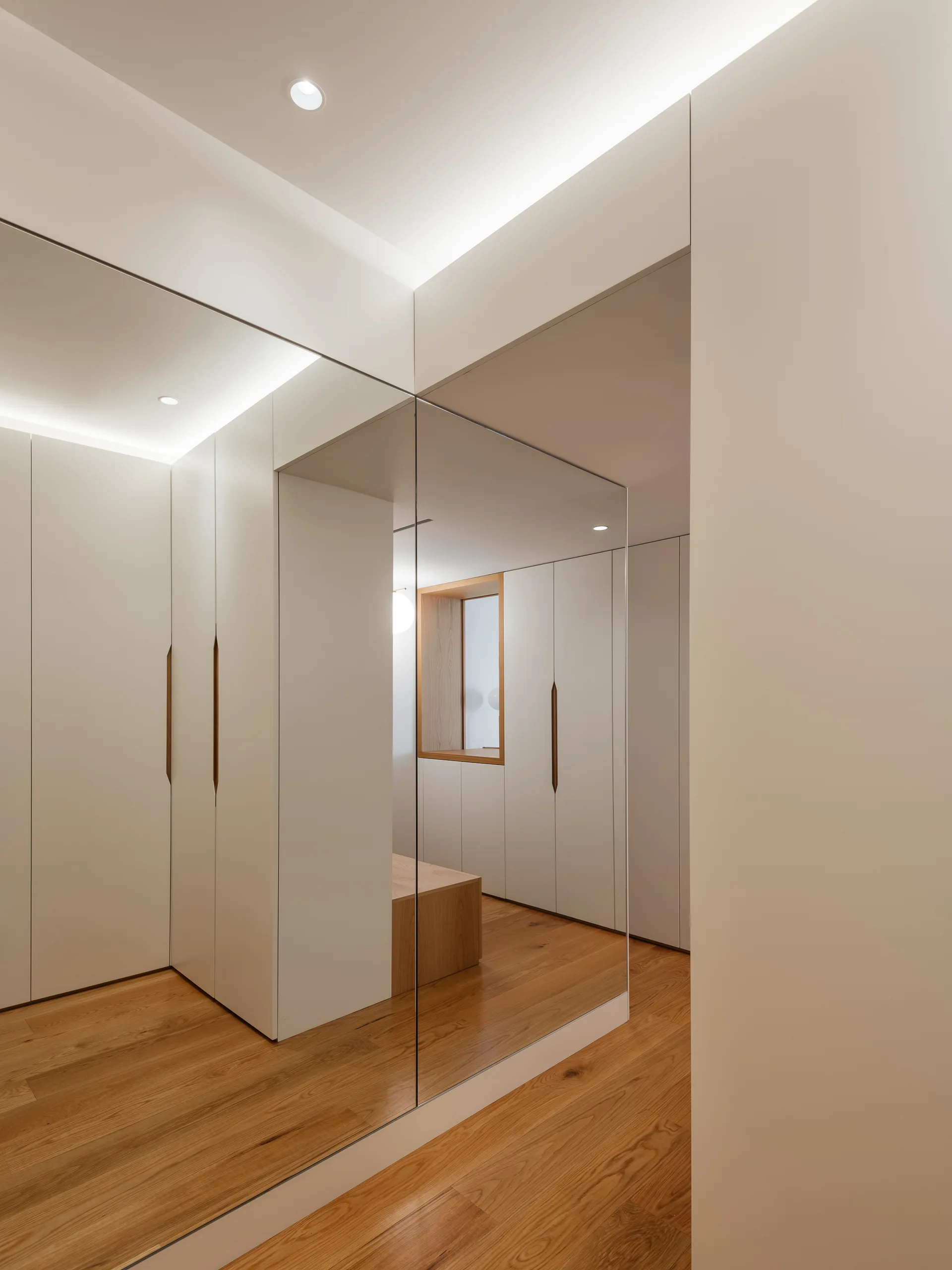 A minimalist white hallway with warm wood floors features built-in cabinets with wood pulls and a large reflective floor-to-ceiling mirror.