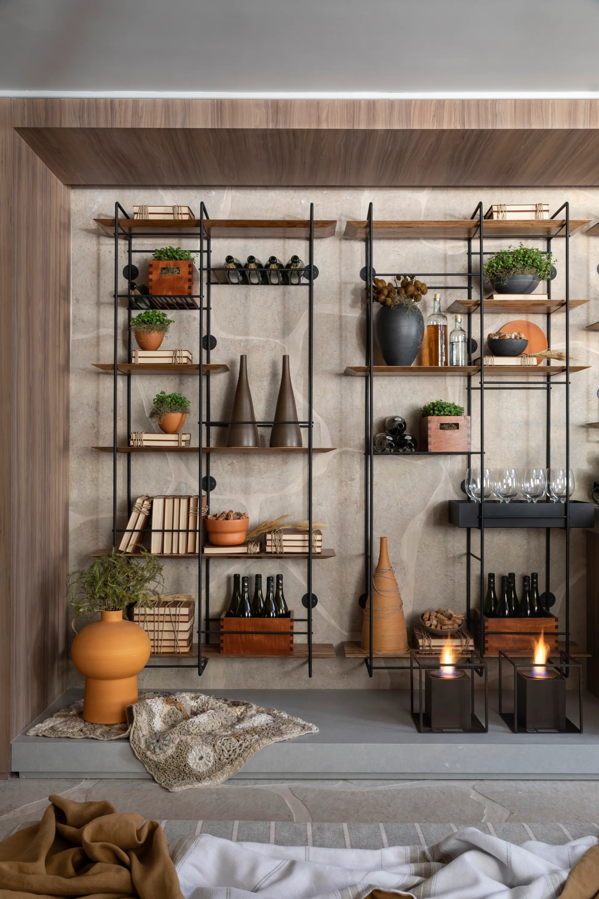 Black metal open shelving holds wine bottles and clay pottery against a textured stone wall within a wood-framed niche.