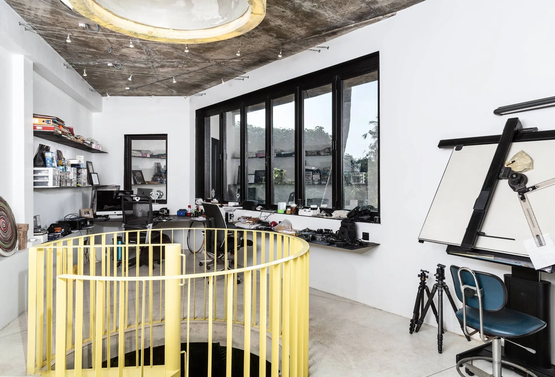 A modern home office features a yellow spiral staircase railing, desks with monitors, large black-frame windows, and an exposed concrete ceiling.