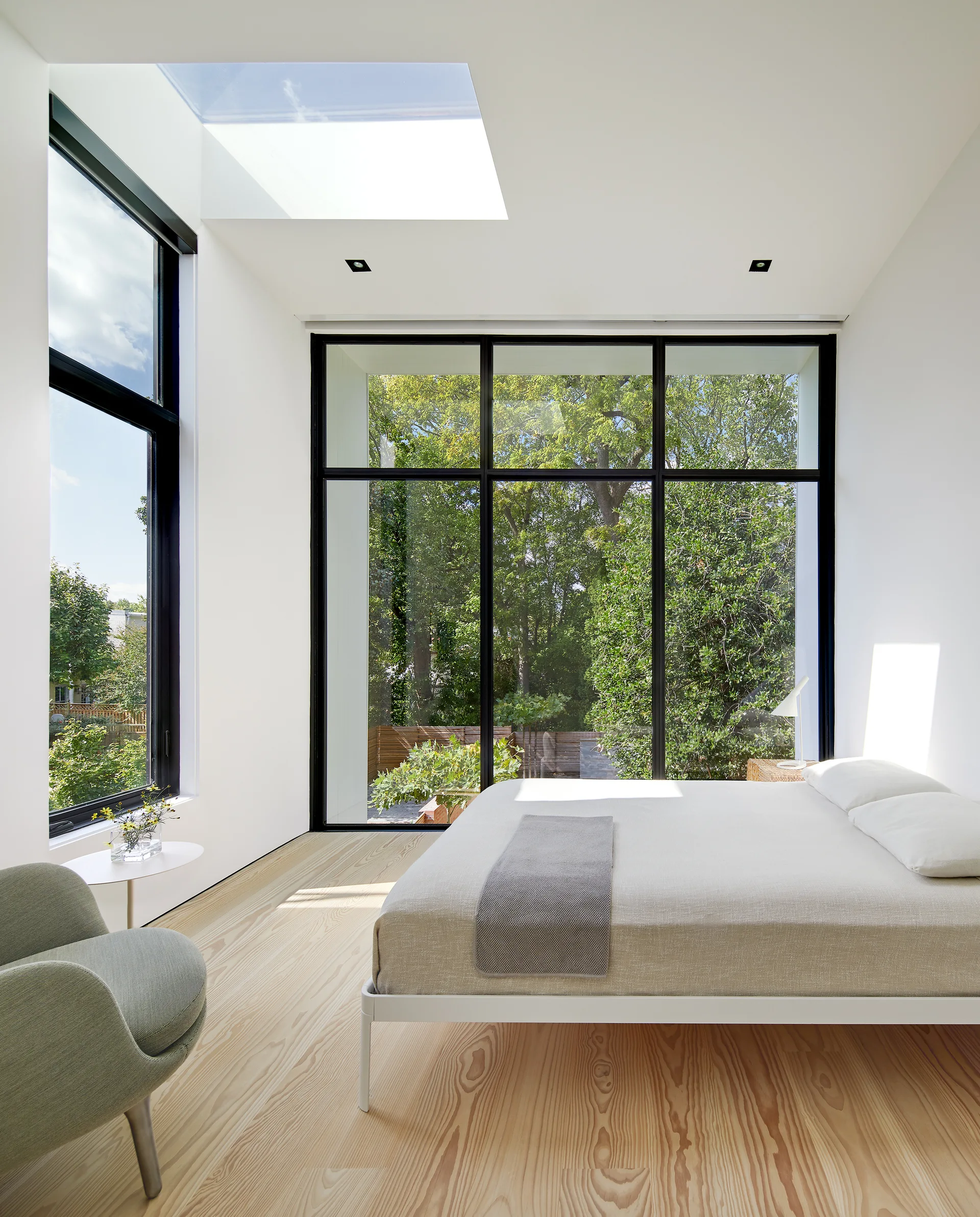 A minimalist bedroom featuring light wood floors, a low-profile white bed, a skylight, and large black-framed windows overlooking green trees.