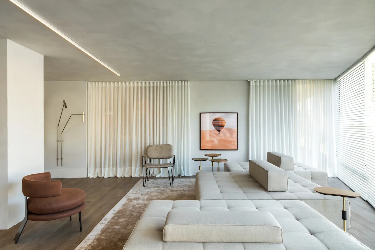 A minimalist living room with a low-profile modular sofa, a rust-colored armchair, and floor-to-ceiling sheer curtains.