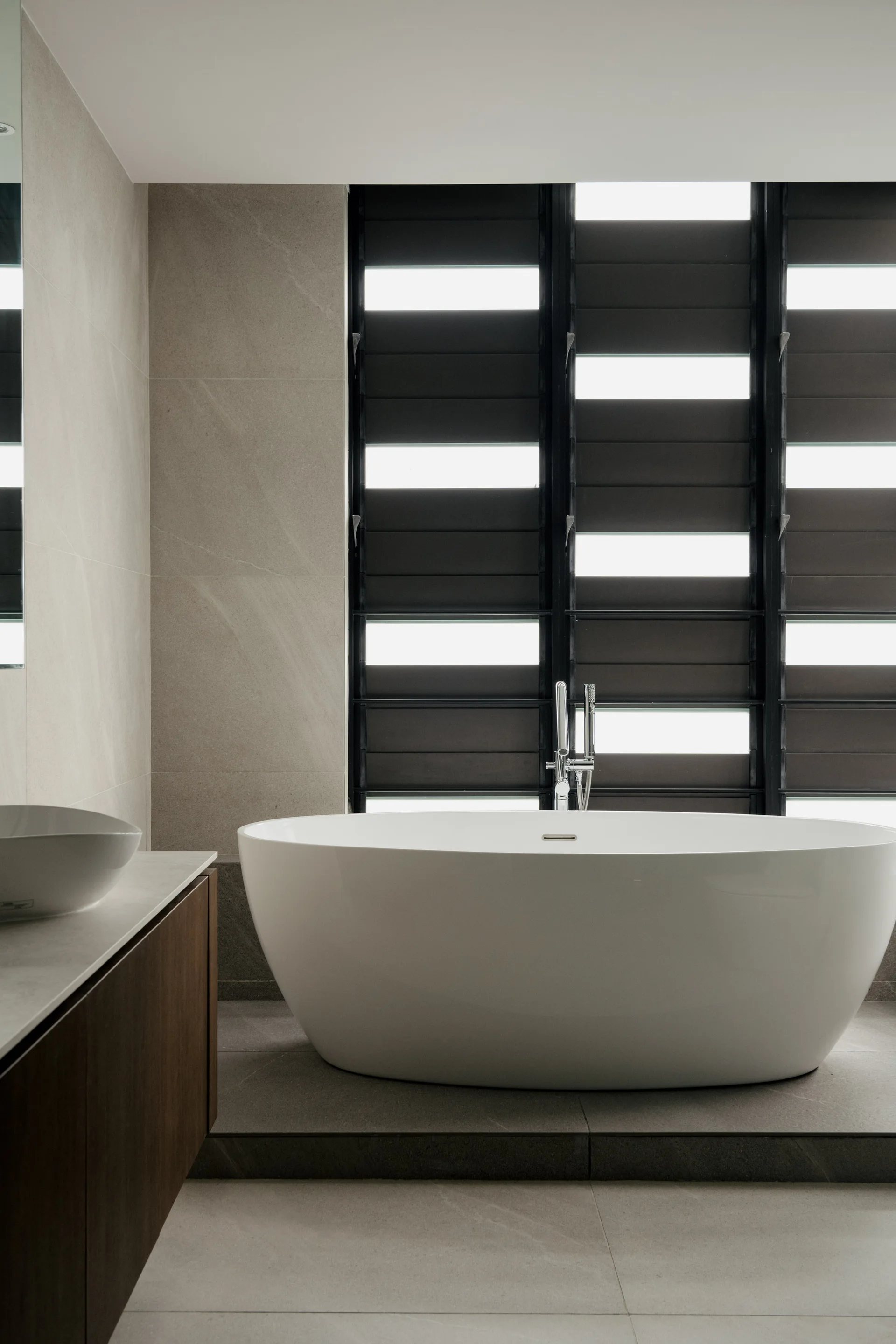 A minimalist bathroom features a white oval freestanding tub in front of black louvered windows and a wood vanity with a white vessel sink.