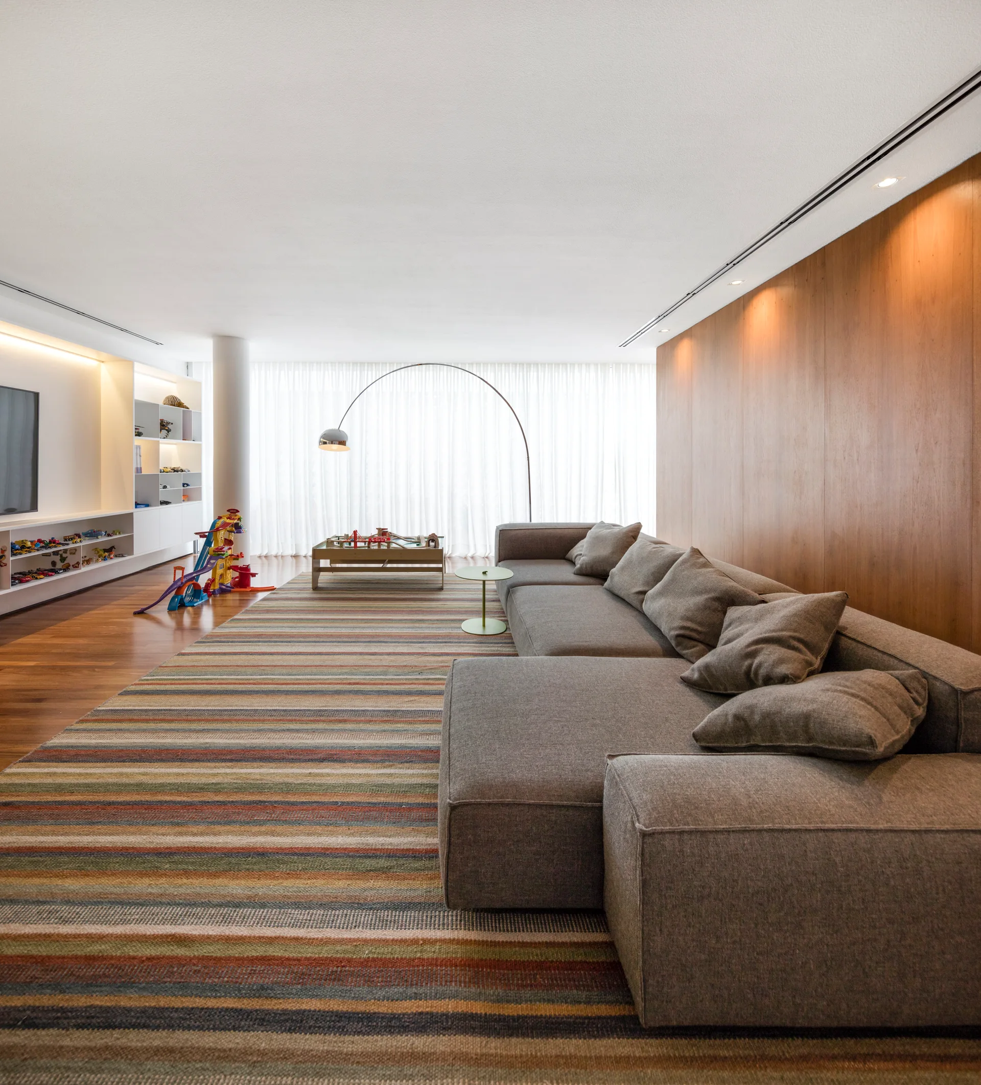 A grey sectional sofa sits on a colorful striped rug in a contemporary living room with a wood-paneled wall and built-in toy shelves.
