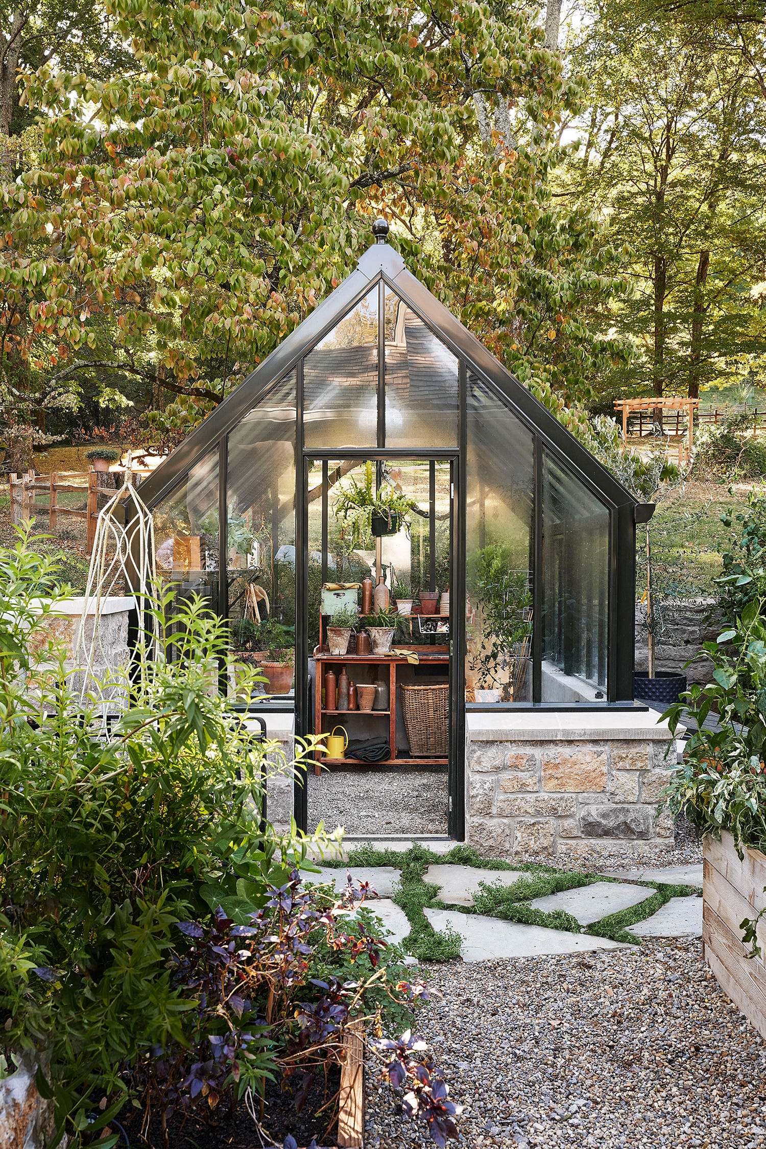 A backyard glass greenhouse with a black metal gabled frame and a natural stone base features a wooden potting bench inside.