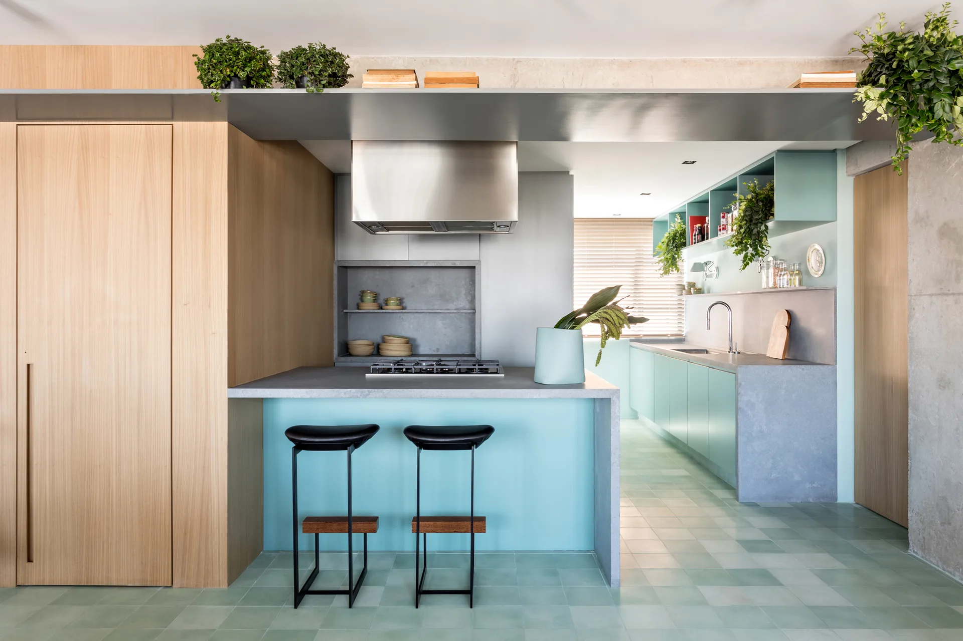 A modern kitchen features a teal island with two black stools, natural wood wall paneling, gray stone countertops, and light green tiled flooring.