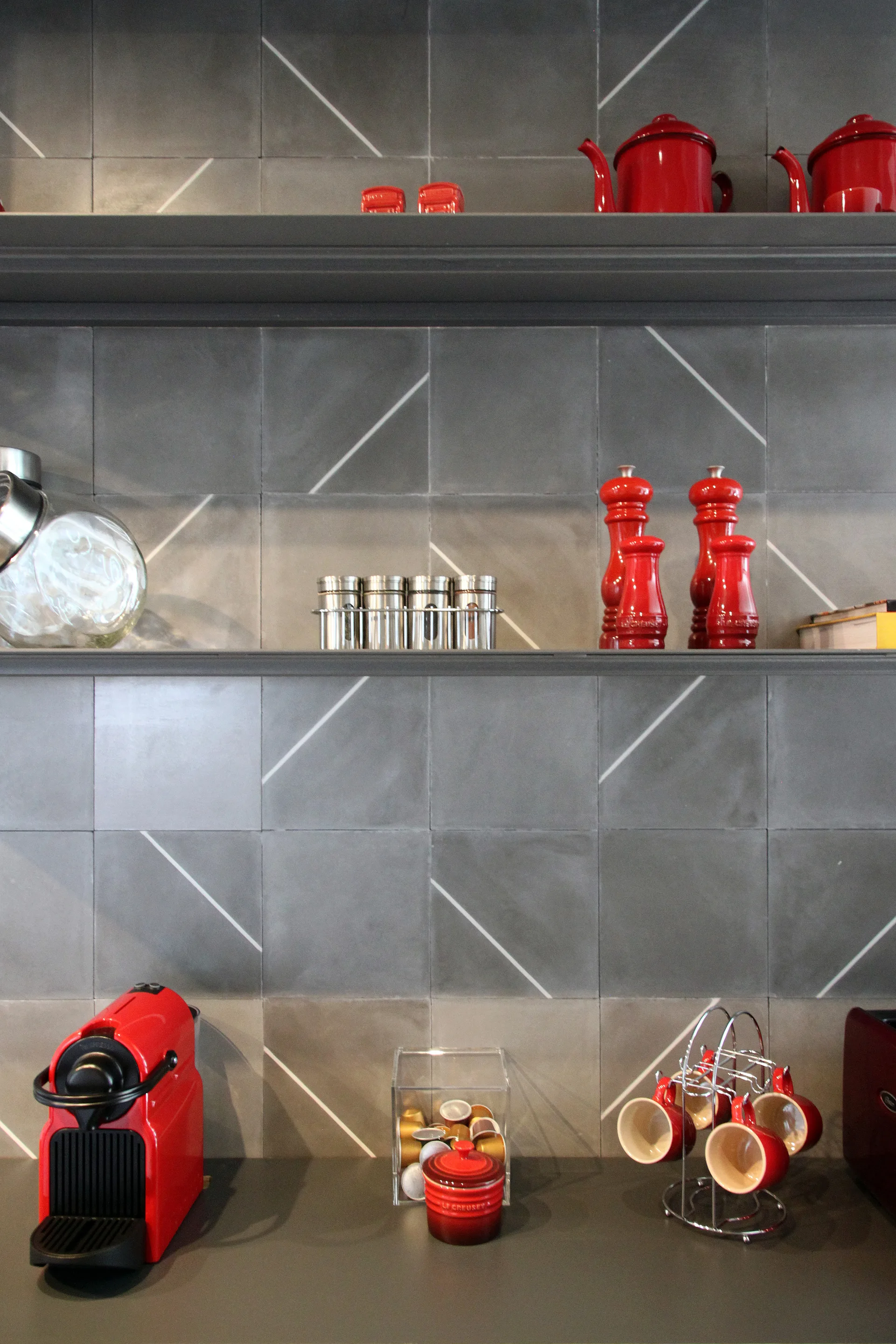 Dark metal open shelves mounted on a grey tiled wall with white diagonal stripes, holding cherry red kitchenware and coffee accessories.