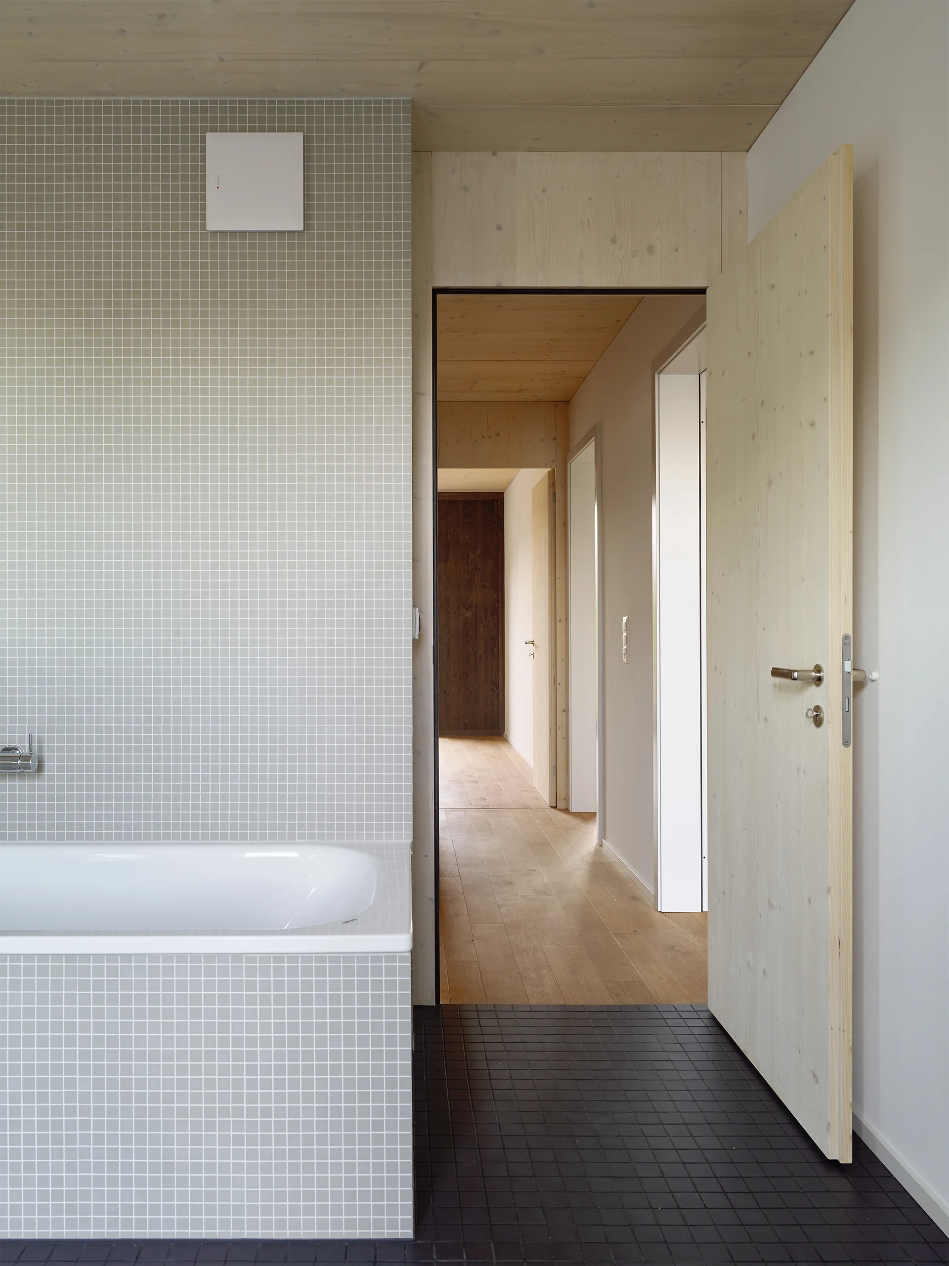 A minimalist bathroom features grey square tile walls, a matching tiled bathtub, black floor tiles, and an open pale wood door leading to a hallway.