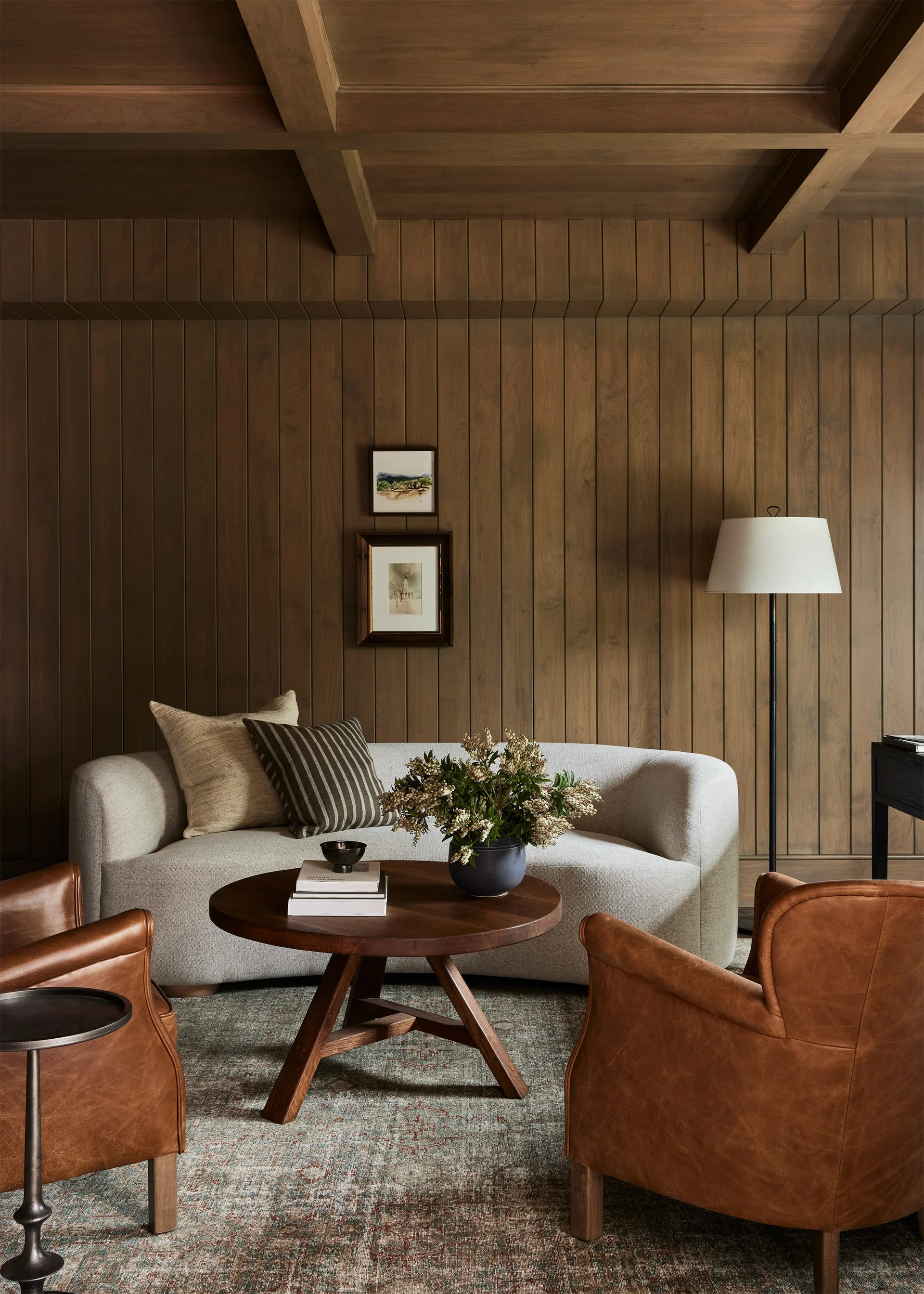 A warm-toned living room features a beige curved sofa, two leather armchairs, and a round wood coffee table against vertical wood paneling.