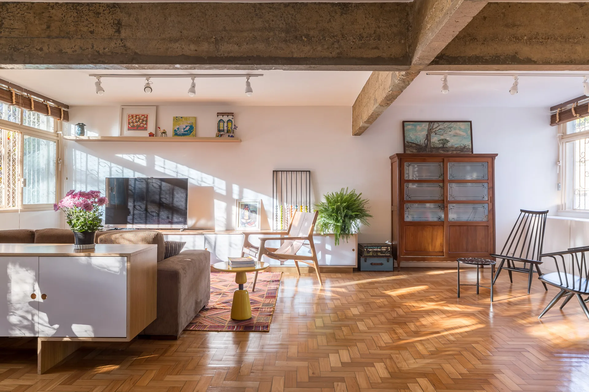 An eclectic living room with herringbone parquet wood floors, an exposed concrete beam, and mid-century furniture under natural sunlight.