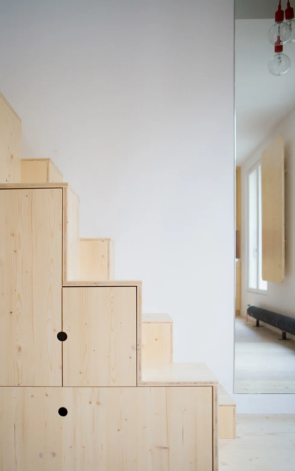 A light wood storage staircase with round cutout handles stands against a white wall next to a tall vertical mirror reflecting a window.