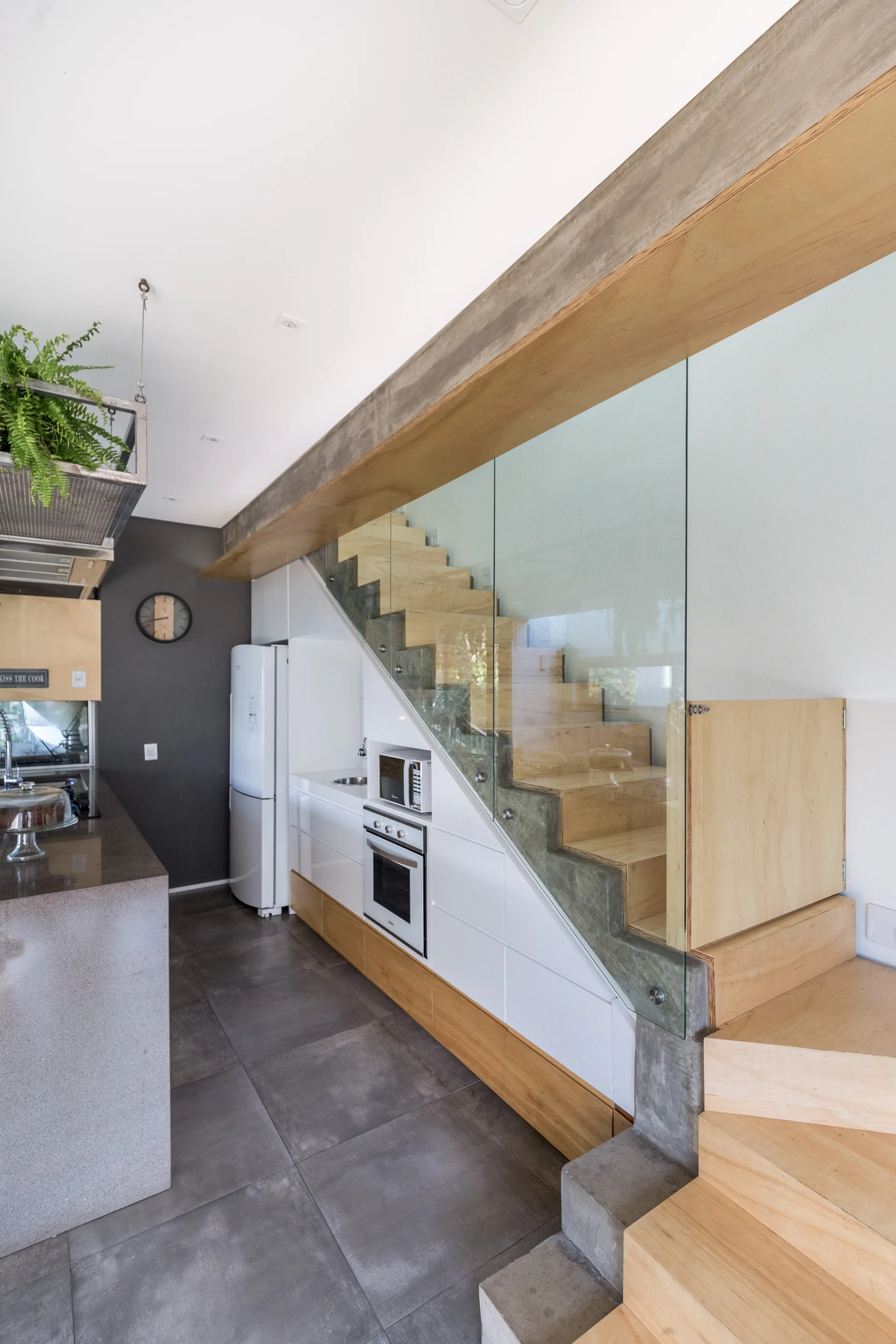 A modern kitchen built directly beneath a light wood staircase featuring a clear glass railing, white and wood cabinetry, and an exposed concrete beam.