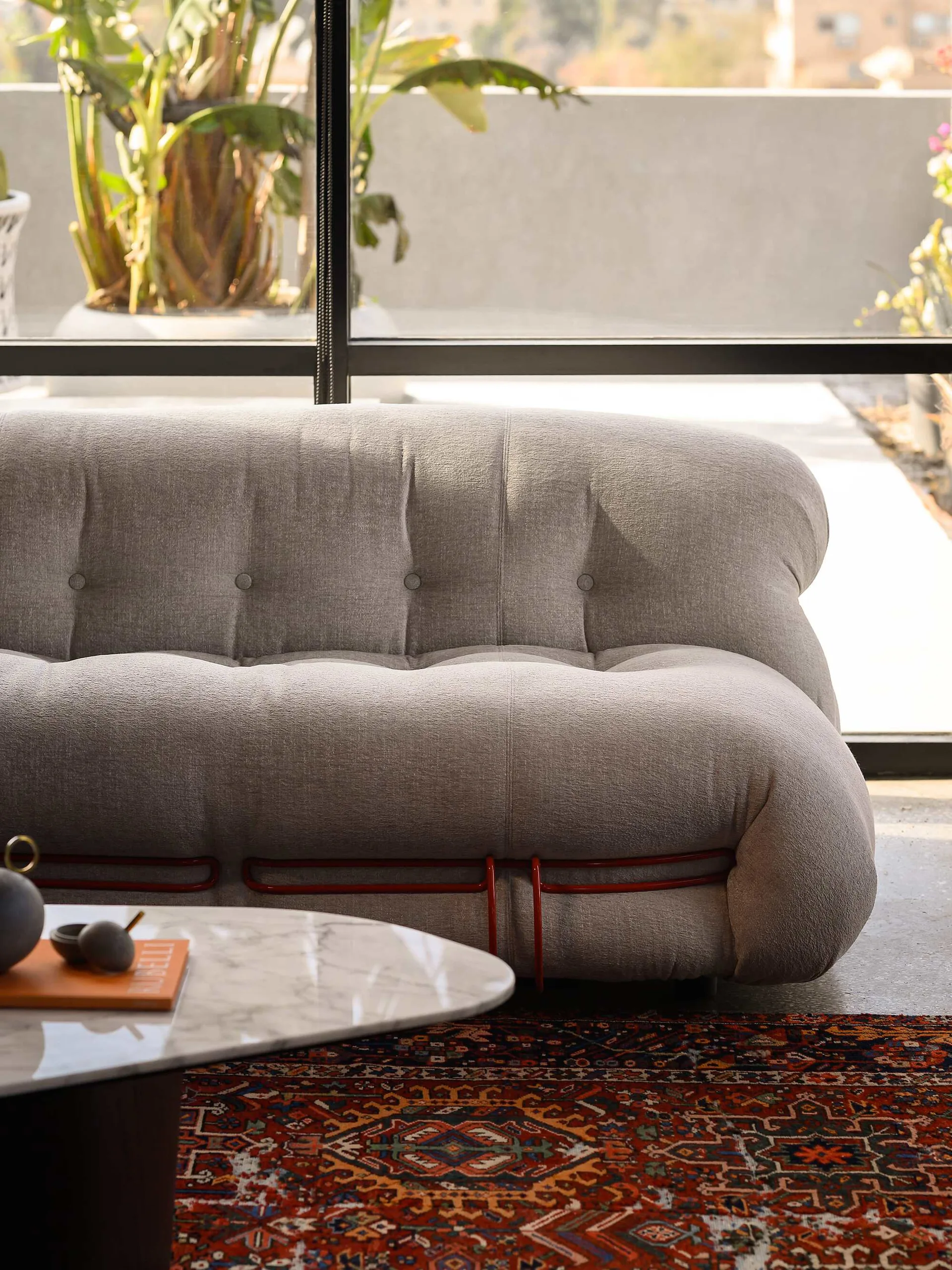 A light gray tufted sofa with a red metal base sits on a patterned area rug next to a marble coffee table in front of a window.