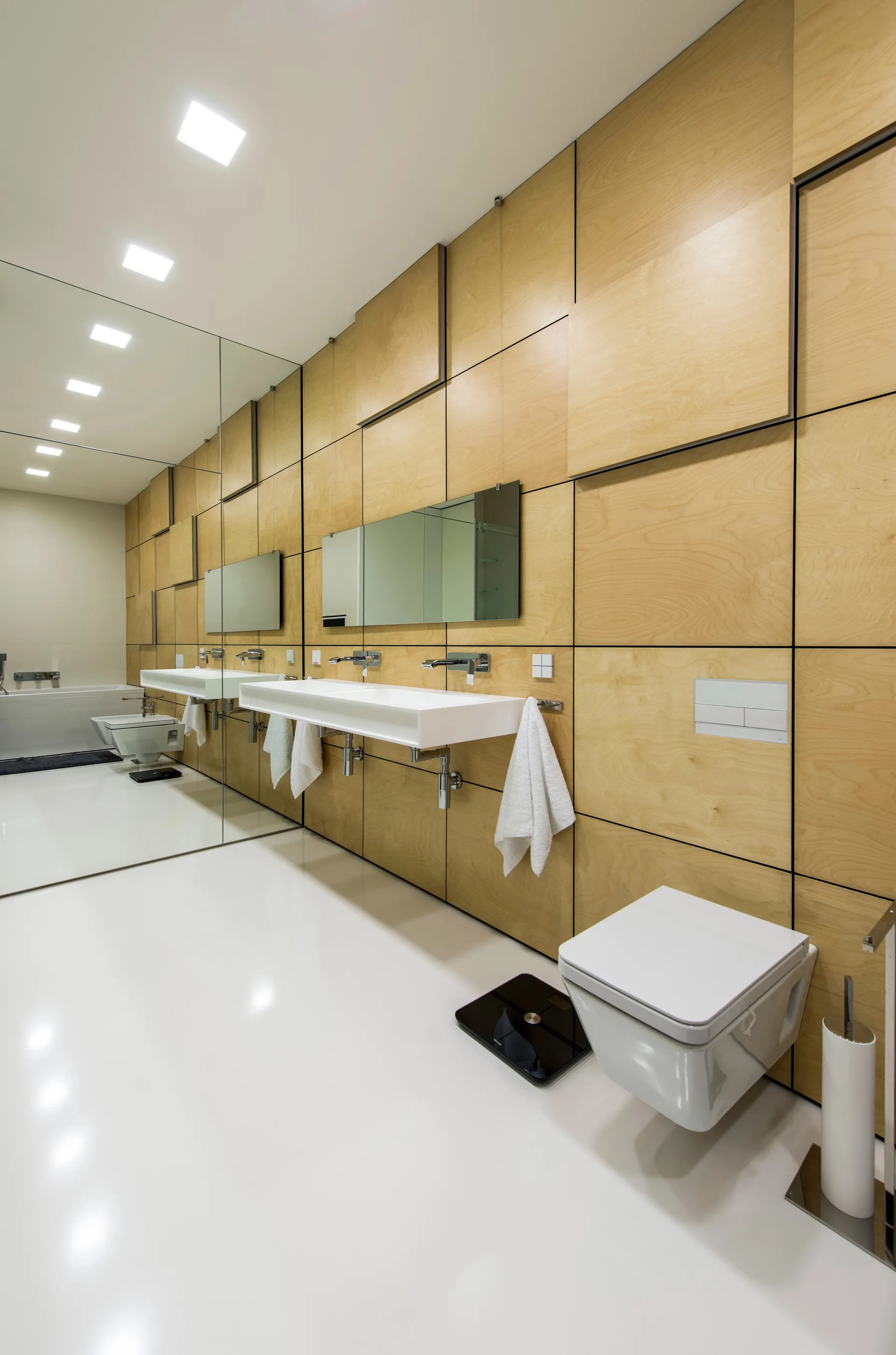 A modern bathroom features light wood paneled walls, a white wall-mounted trough sink, a wall-mounted toilet, and a large mirrored wall reflecting the space.
