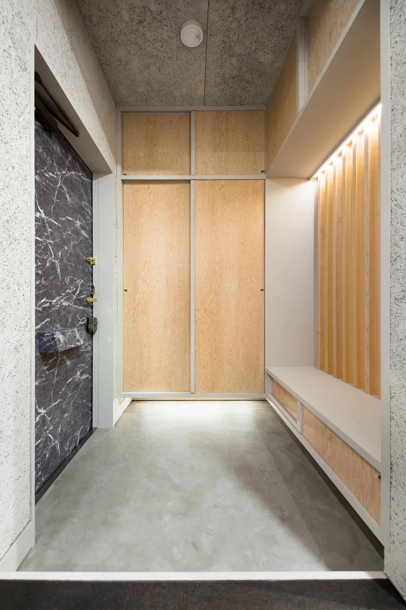 A minimalist entryway features built-in plywood sliding wardrobes, a concrete floor, and an integrated bench with a slatted wood panel.