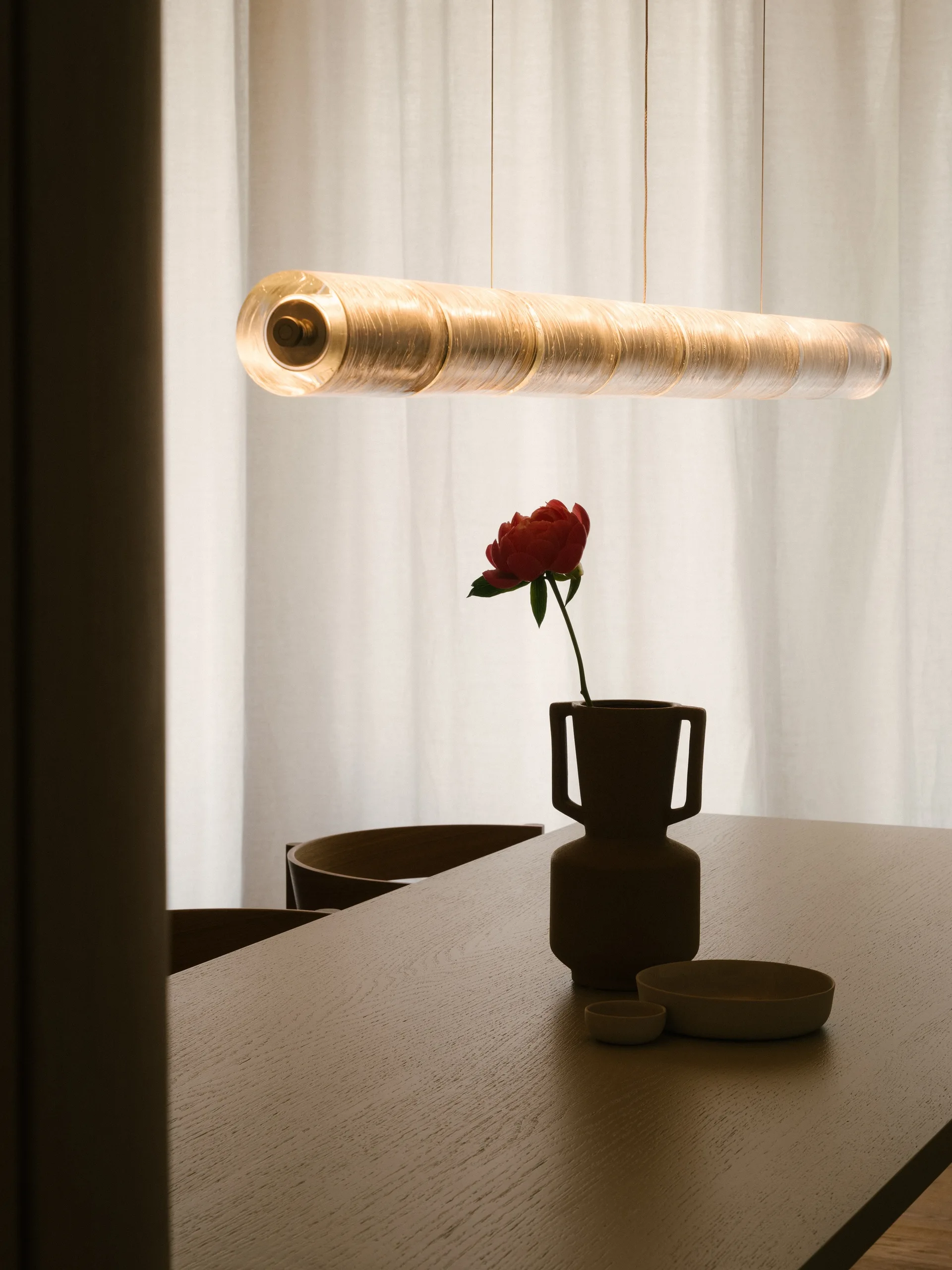 A wooden dining table features a small ceramic vase with a single red flower beneath a glowing cylindrical glass pendant light.